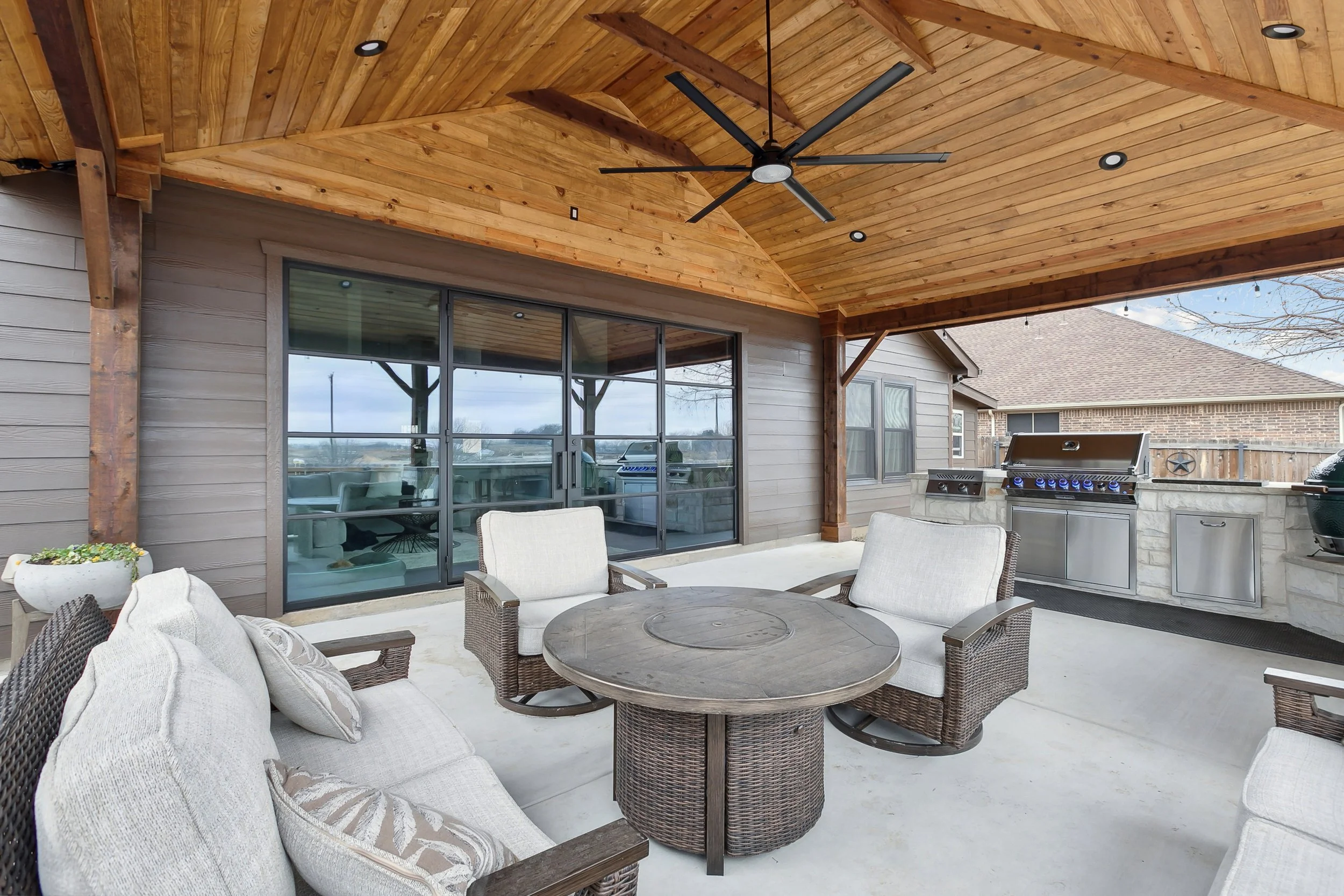 An outdoor patio with a wooden ceiling, ceiling fan, and rectangular table with wicker chairs, overlooking a backyard with a grill and a neighboring brick house.