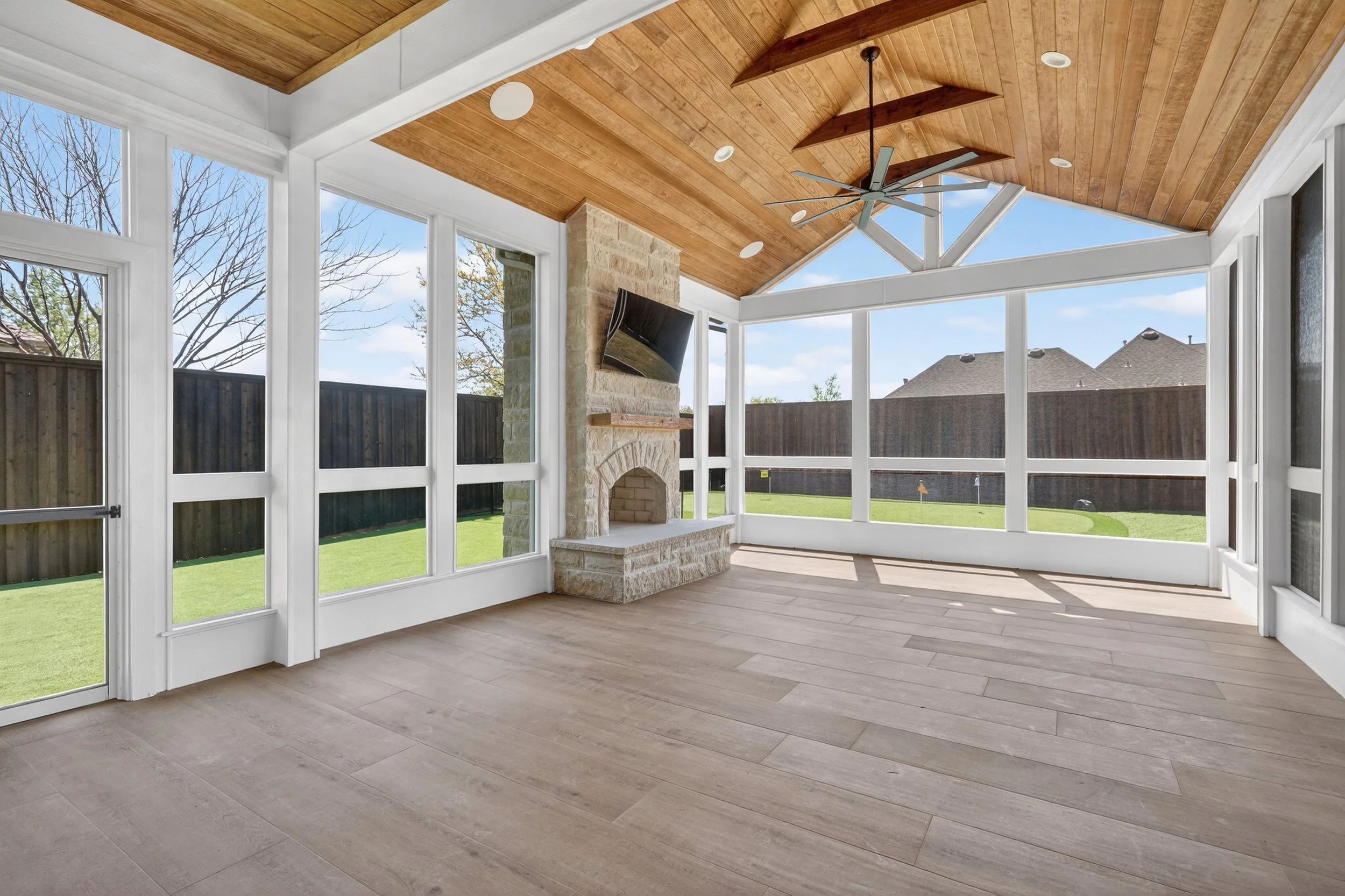 Sunroom with large windows, overlooking a backyard with a wooden fence, trees, and houses with sloped roofs. The ceiling is wood-paneled with a ceiling fan and recessed lights. The stone fireplace is on one wall.