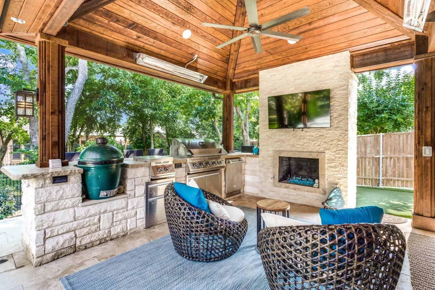 An outdoor covered patio with a wooden ceiling, ceiling fan, and a stone fireplace with a TV mounted above it. There is a built-in grill and a green egg smoker on a stone counter, surrounded by trees and a fenced yard. Two wicker chairs with blue cus