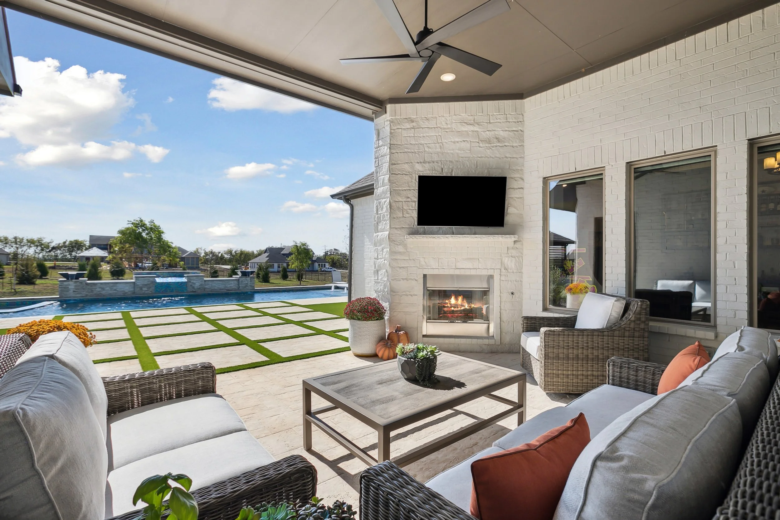 Outdoor patio with white cushioned wicker furniture, a coffee table with plants, a brick fireplace, a TV mounted above the fireplace, decorations including pumpkins and flowers, a sliding glass door, and a view of a pool, yard, and houses under a blu
