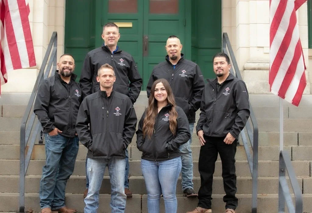 Seven people, six men and one woman, standing on steps outside a building with green doors, wearing black jackets with a logo, posing for a group photo.