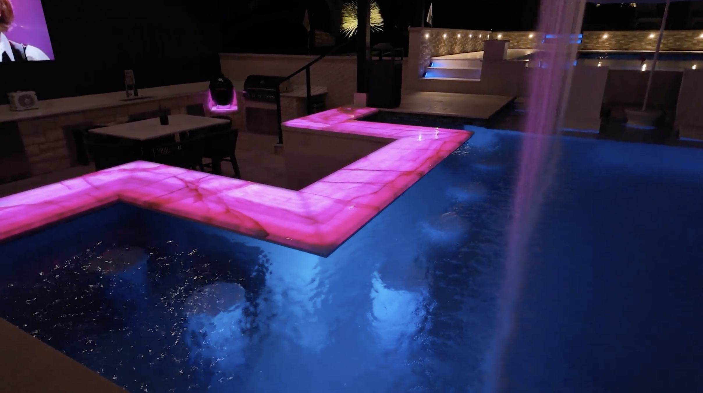Nighttime scene of a pool area with illuminated pink and blue lights. There is a small waterfall feature on the right, and water flows into the pool. Surrounding area has seating and decorative lighting.