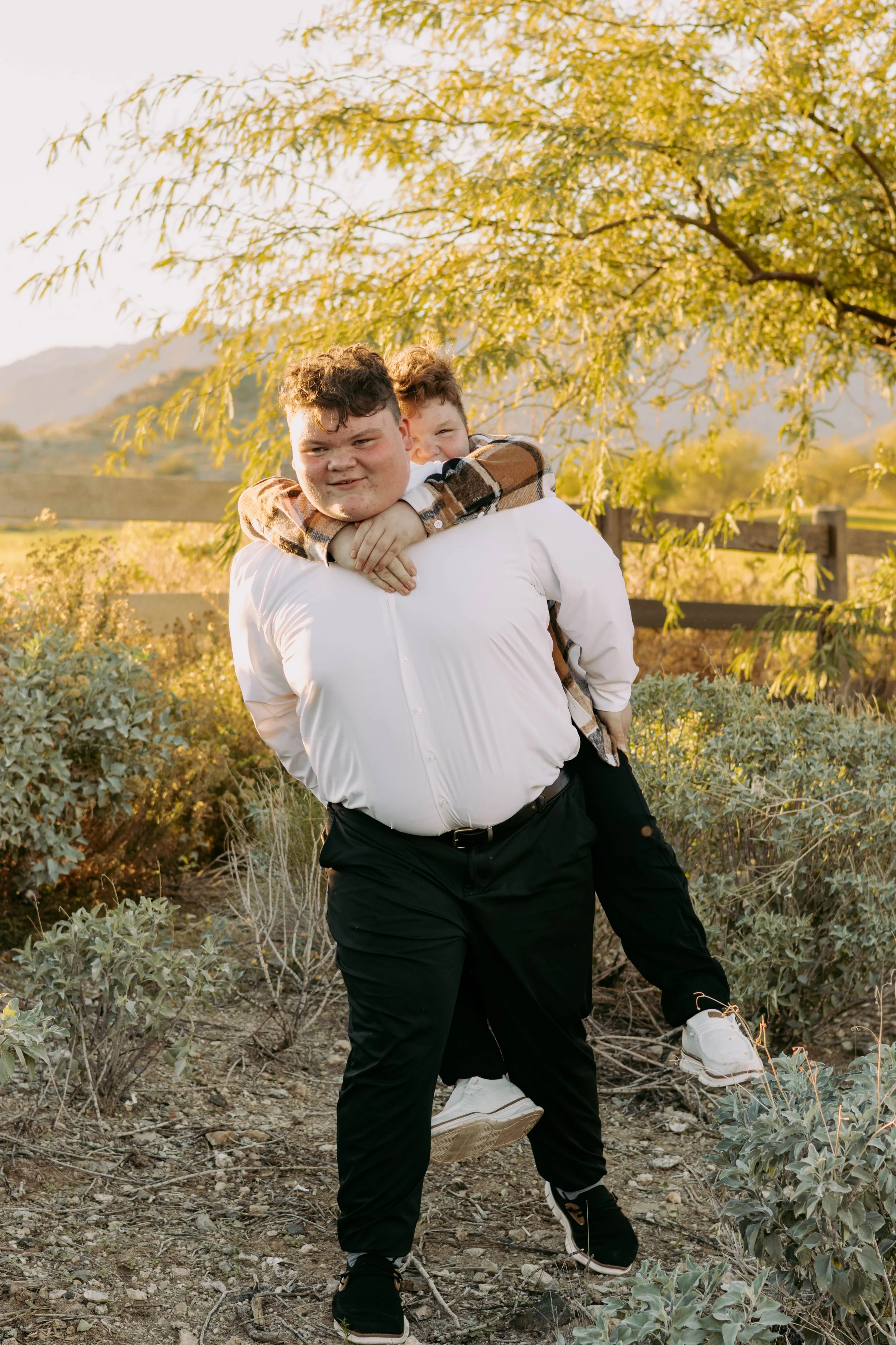 Two young boys outdoors under a tree during sunset, with one boy carrying the other on his back, both smiling and enjoying the moment.