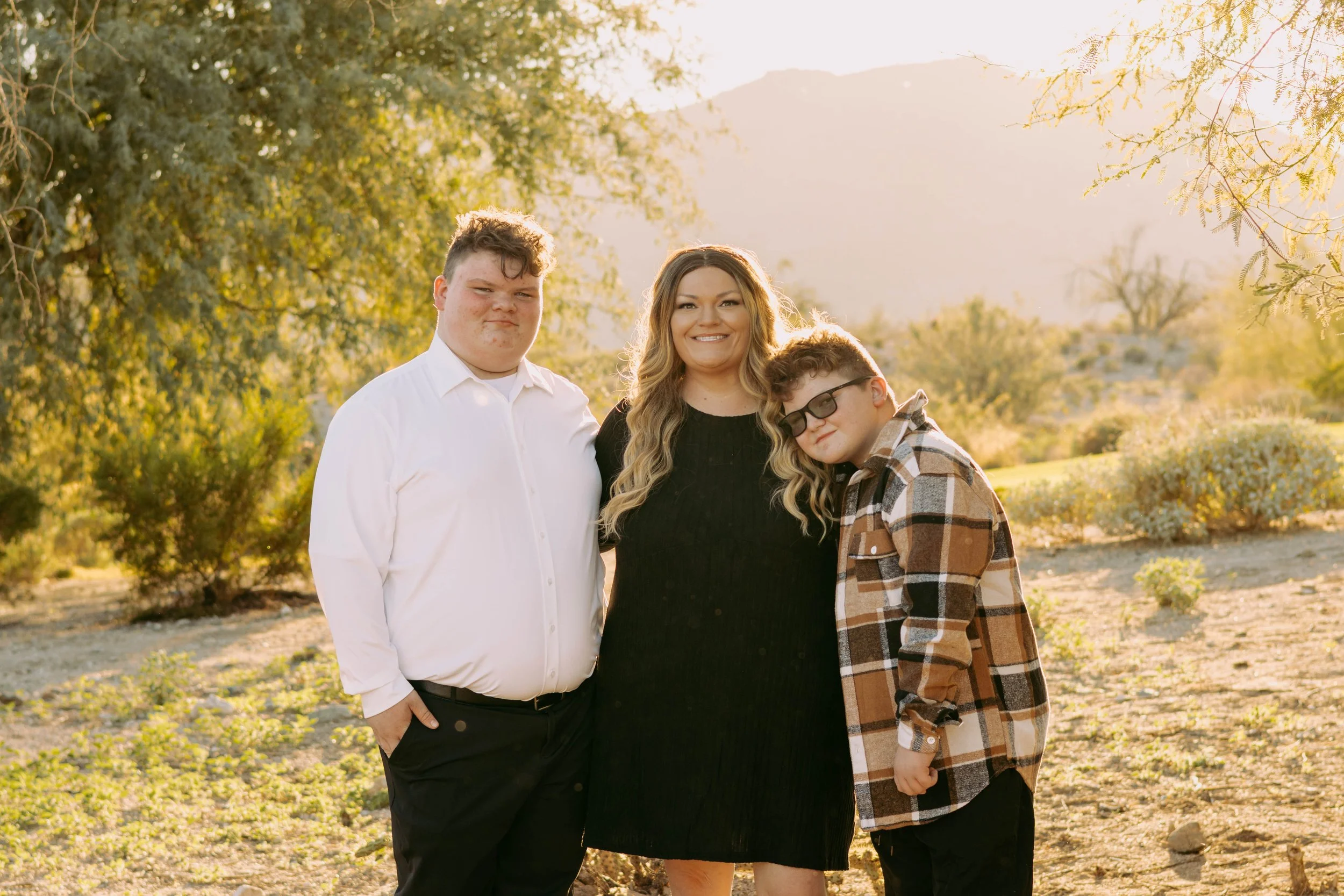 A woman and two boys standing outdoors in a sunny, desert-like area with trees and hills in the background. The woman is smiling, and one boy is wearing glasses and a plaid shirt, while the other boy is wearing a white shirt.