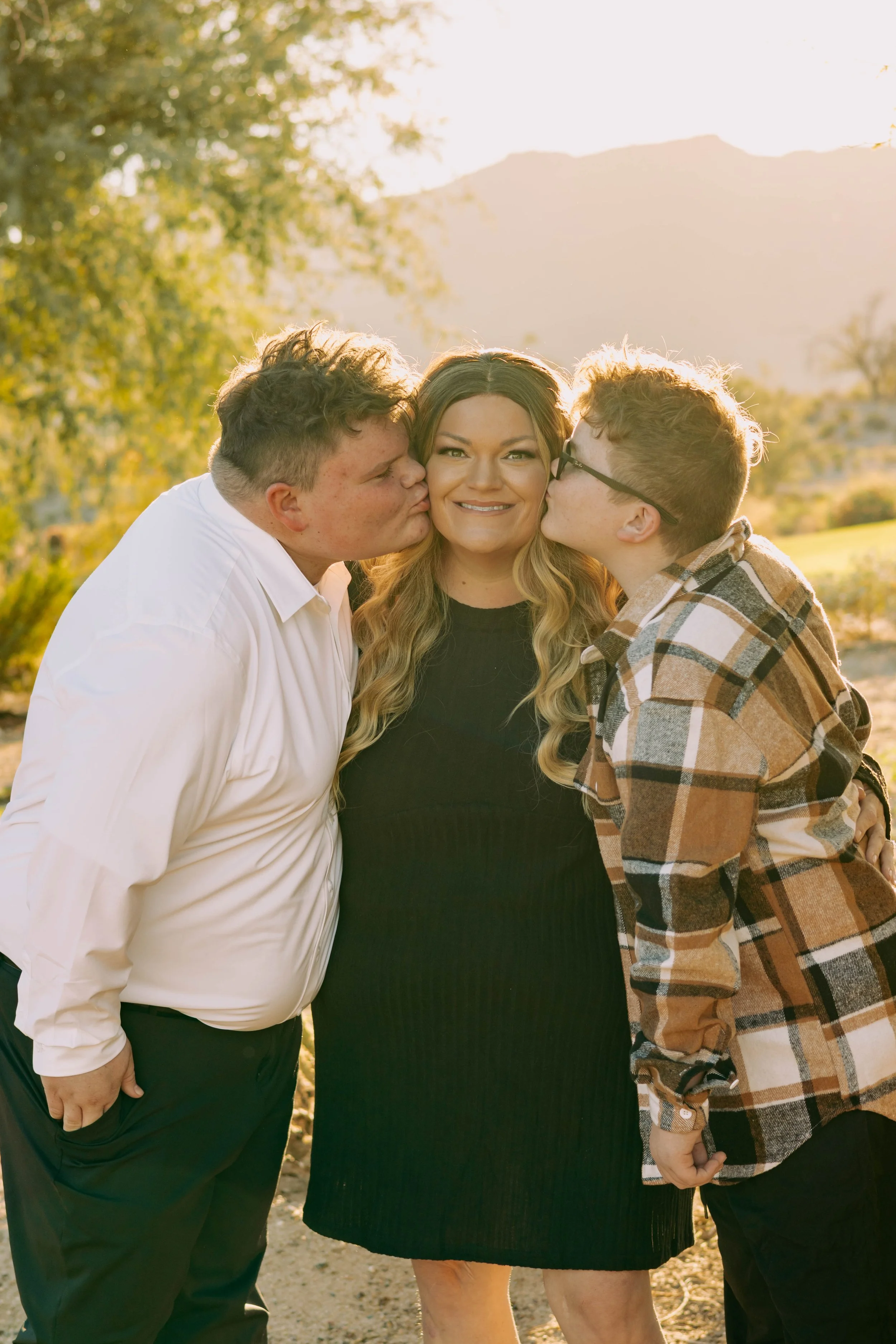 Two boys kiss a smiling woman on each cheek in a park with mountains and trees in the background, during golden hour.