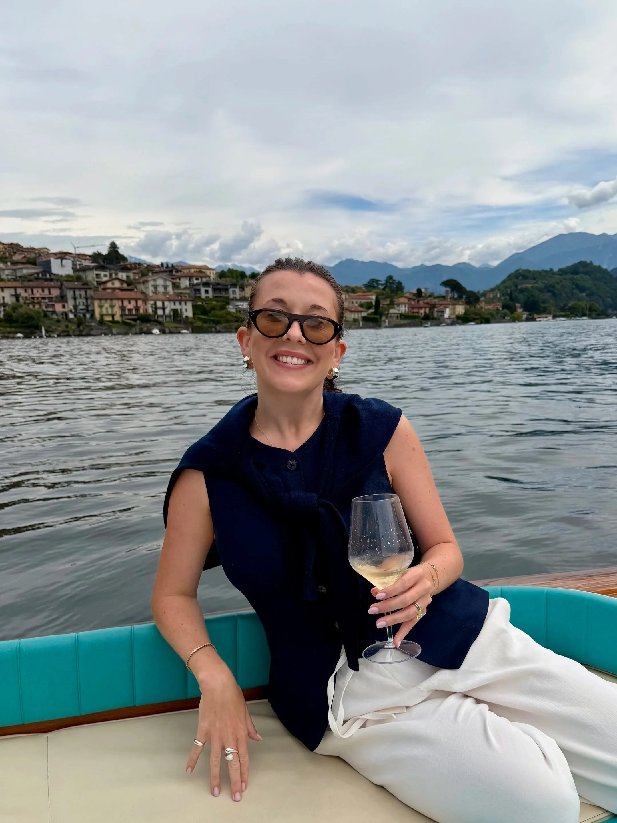 A woman with sunglasses and earrings smiling while sitting on a boat, holding a glass of white wine, with a lake and houses in the background and mountains and clouds in the distance.