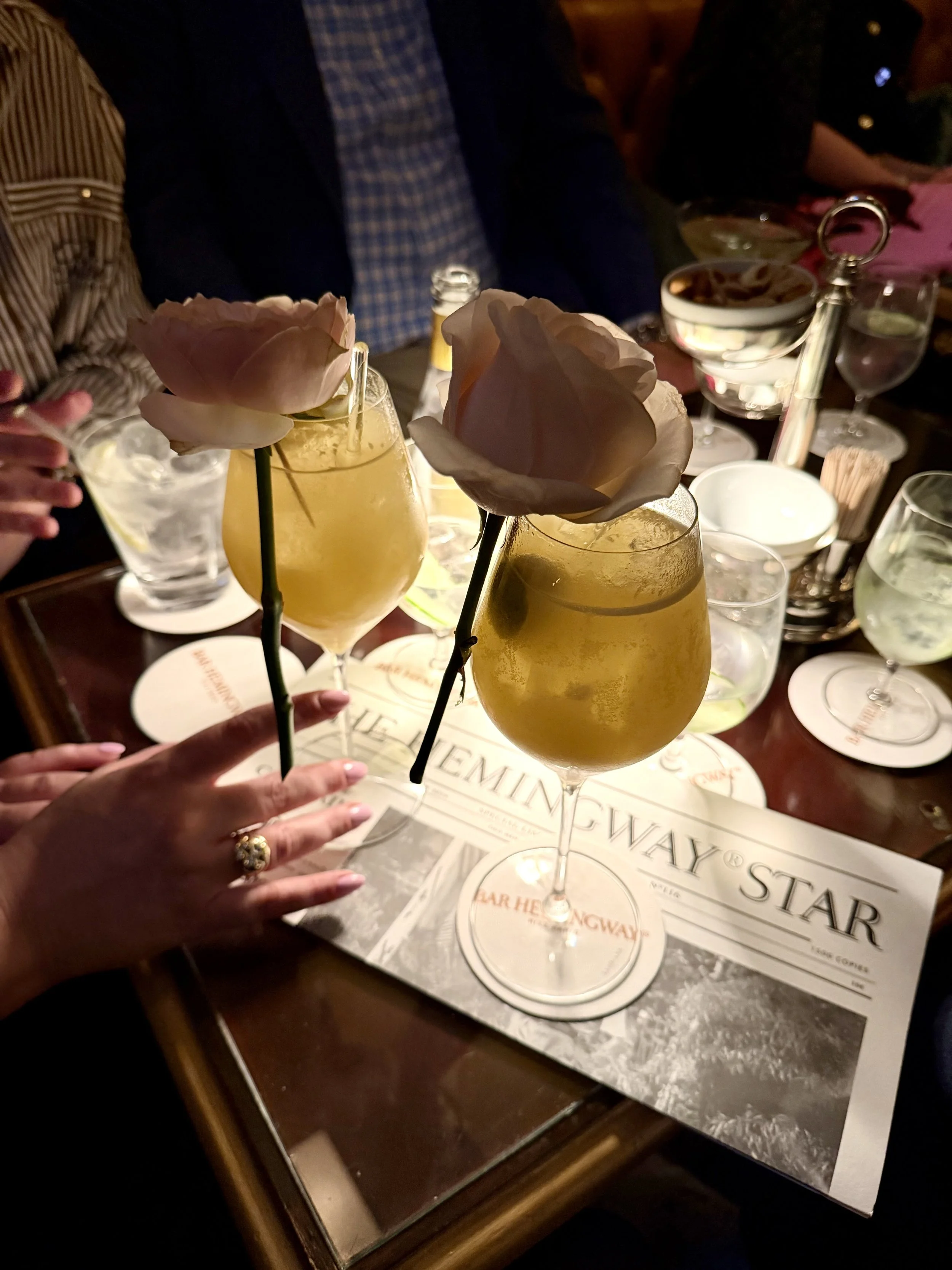 A group of people gather around a table sharing drinks and snacks, with three cocktail glasses topped with pink roses and a menu on the table.