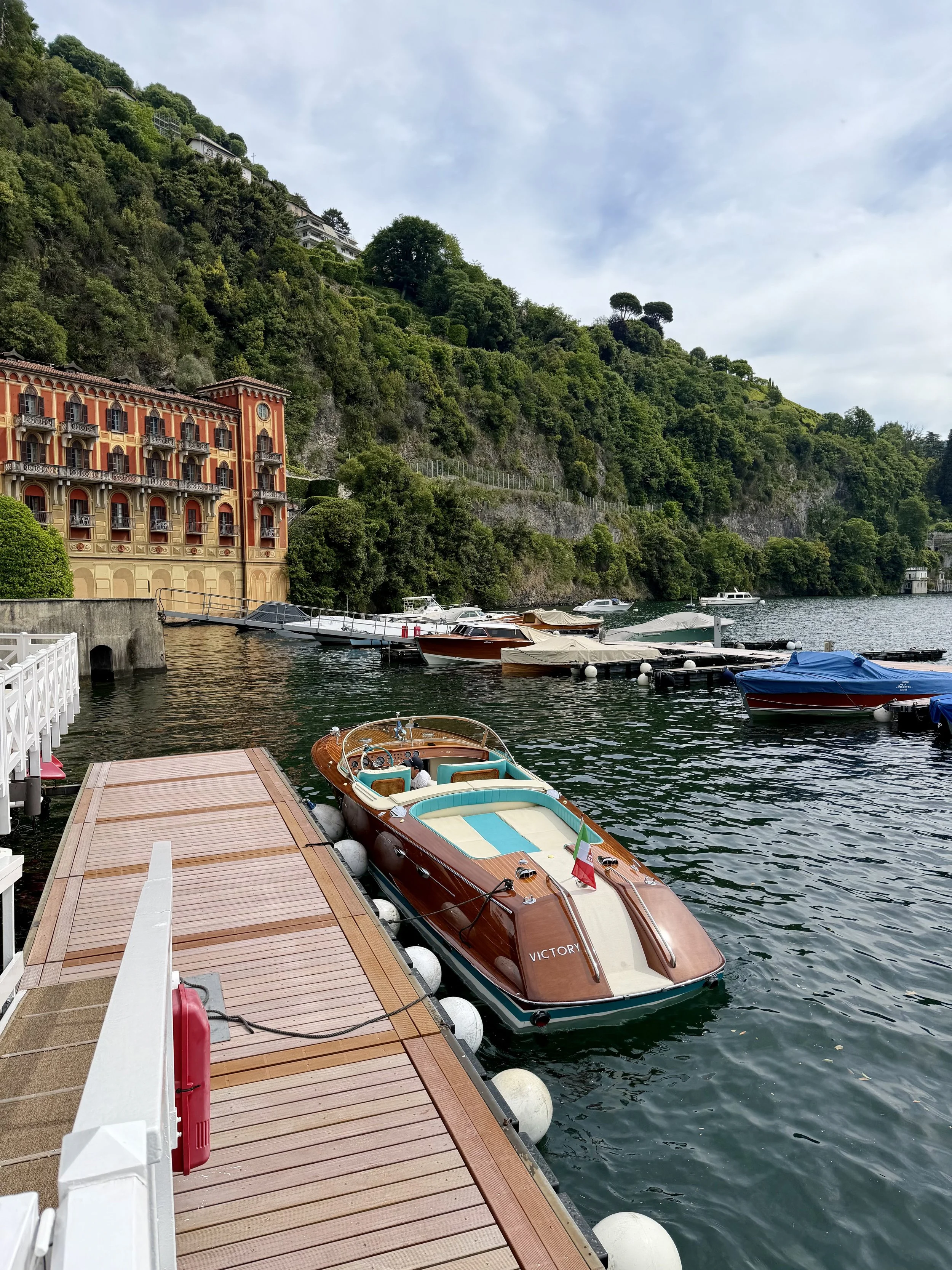 Dock with boats tied to it on a lake, surrounded by green hillside with buildings and trees.