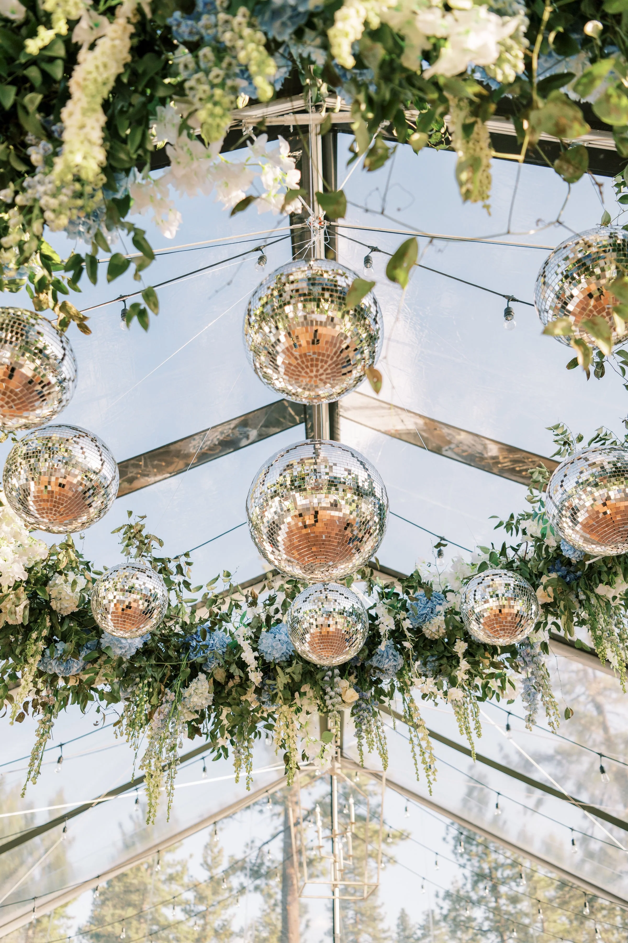 Disco ball decorations hanging from a ceiling surrounded by green leaves and white flowers, with sunlight shining through glass ceiling.