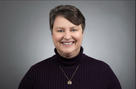 Portrait of a smiling woman with short dark hair, wearing a black turtleneck and a gold necklace, against a gray background.