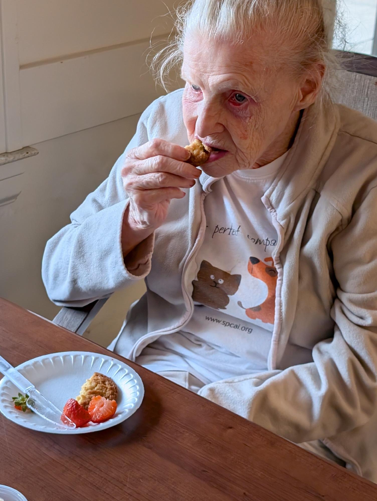 An elderly woman with white hair and wrinkled skin sitting at a wooden table, wearing a beige jacket and a white T-shirt with a bear graphic, eating a piece of food. There are plates of food with strawberries and other items on the table in front of 