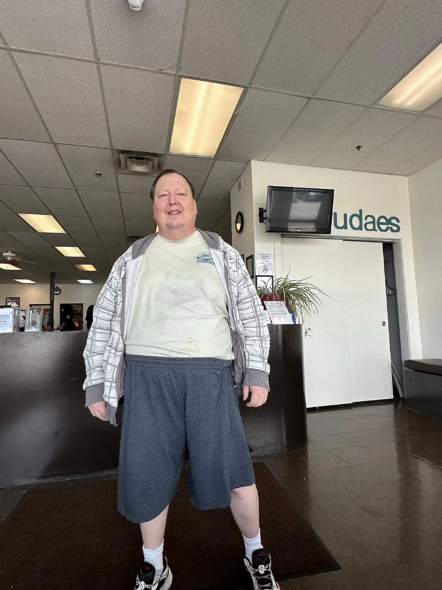 A man standing inside a building near a reception desk with a partial sign that reads "udaes." He is wearing a white T-shirt, gray shorts, a gray hoodie, white socks, and black and white sneakers. The ceiling has fluorescent lights and a dropped ceil