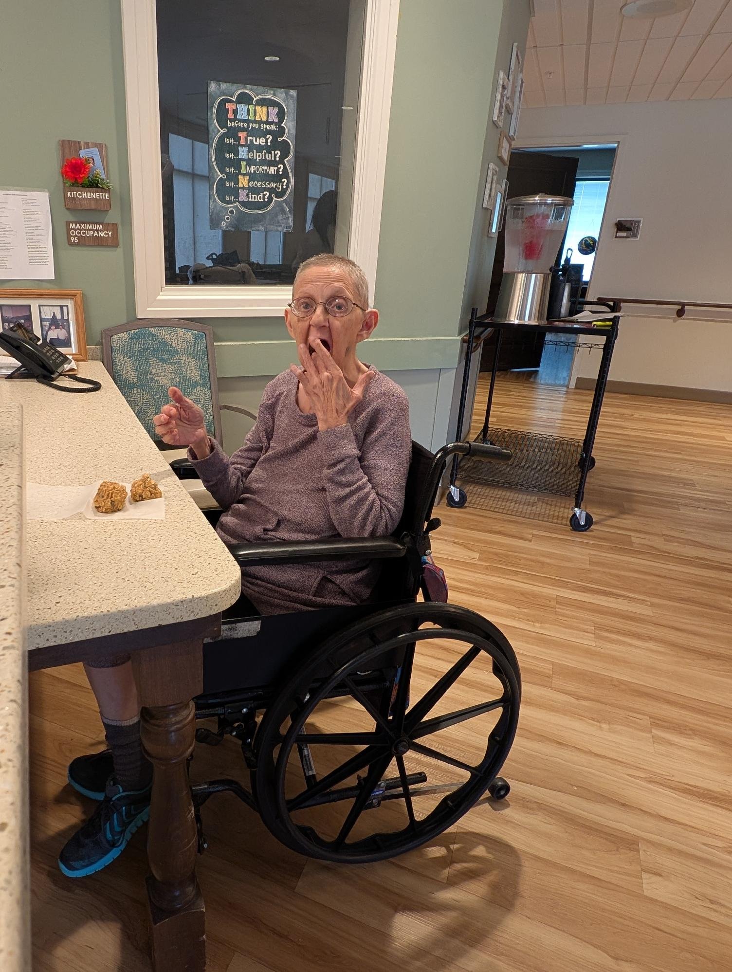 An elderly woman with glasses sitting in a wheelchair at a table, yawning while holding her hand over her mouth. There are two pieces of fried chicken on a napkin in front of her. The background features a wall with a window and a poster, a cart with