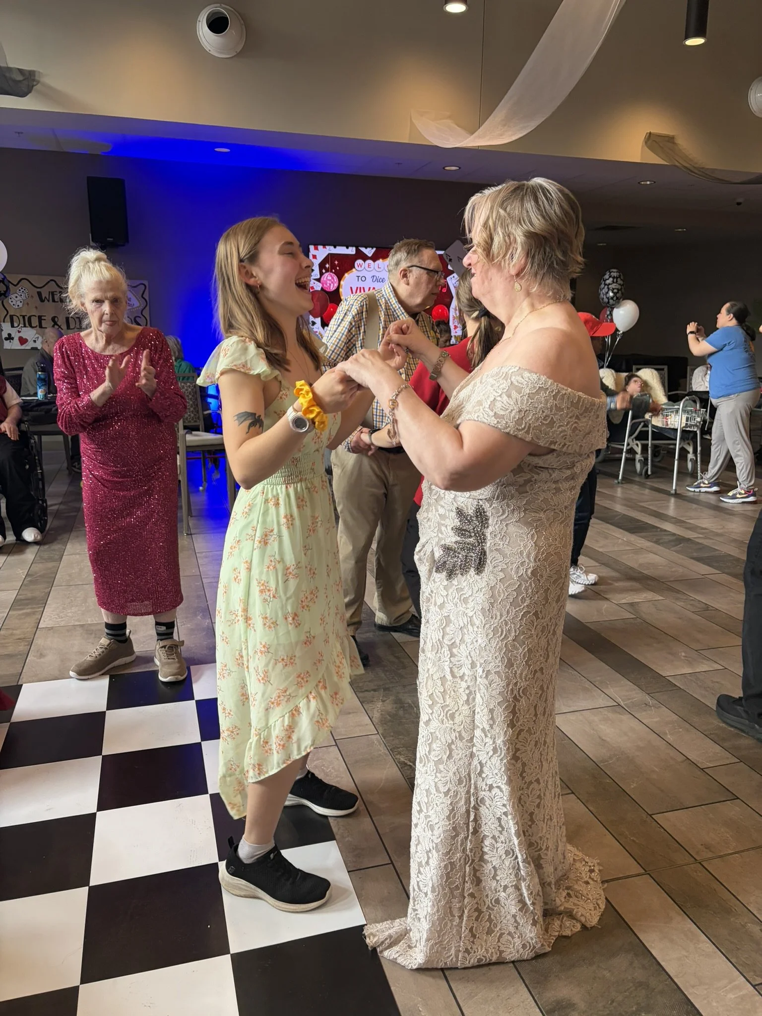A young woman and an older woman dance together, laughing and holding hands, at a festive event with balloons and decorations. Several other people are in the background, some clapping and observing the scene.