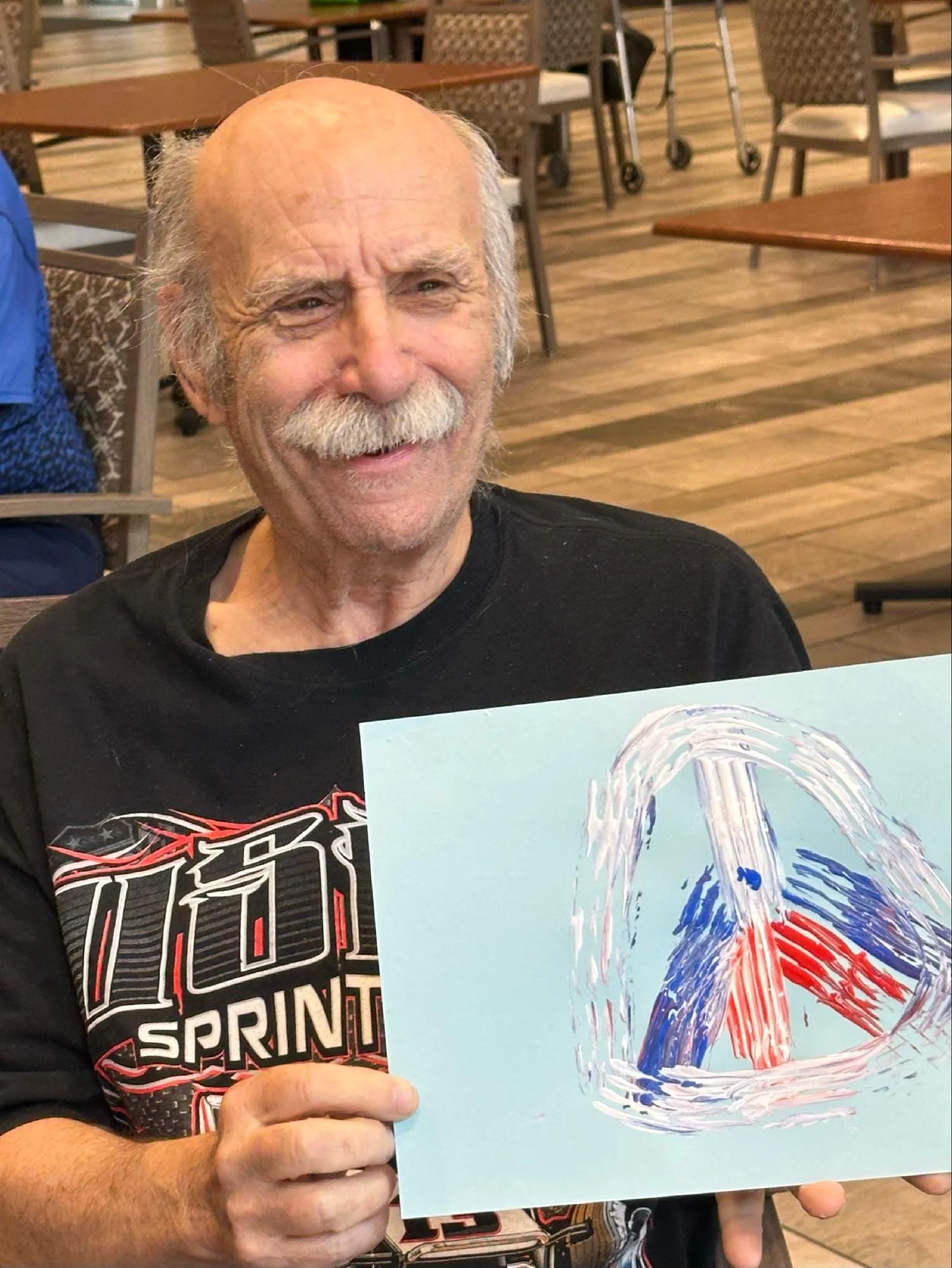 An elderly man with gray hair and a mustache is smiling and holding a piece of artwork depicting a water fountain with colorful water streams on a light blue background. He is seated in a restaurant or cafe with wooden flooring and chairs in the back
