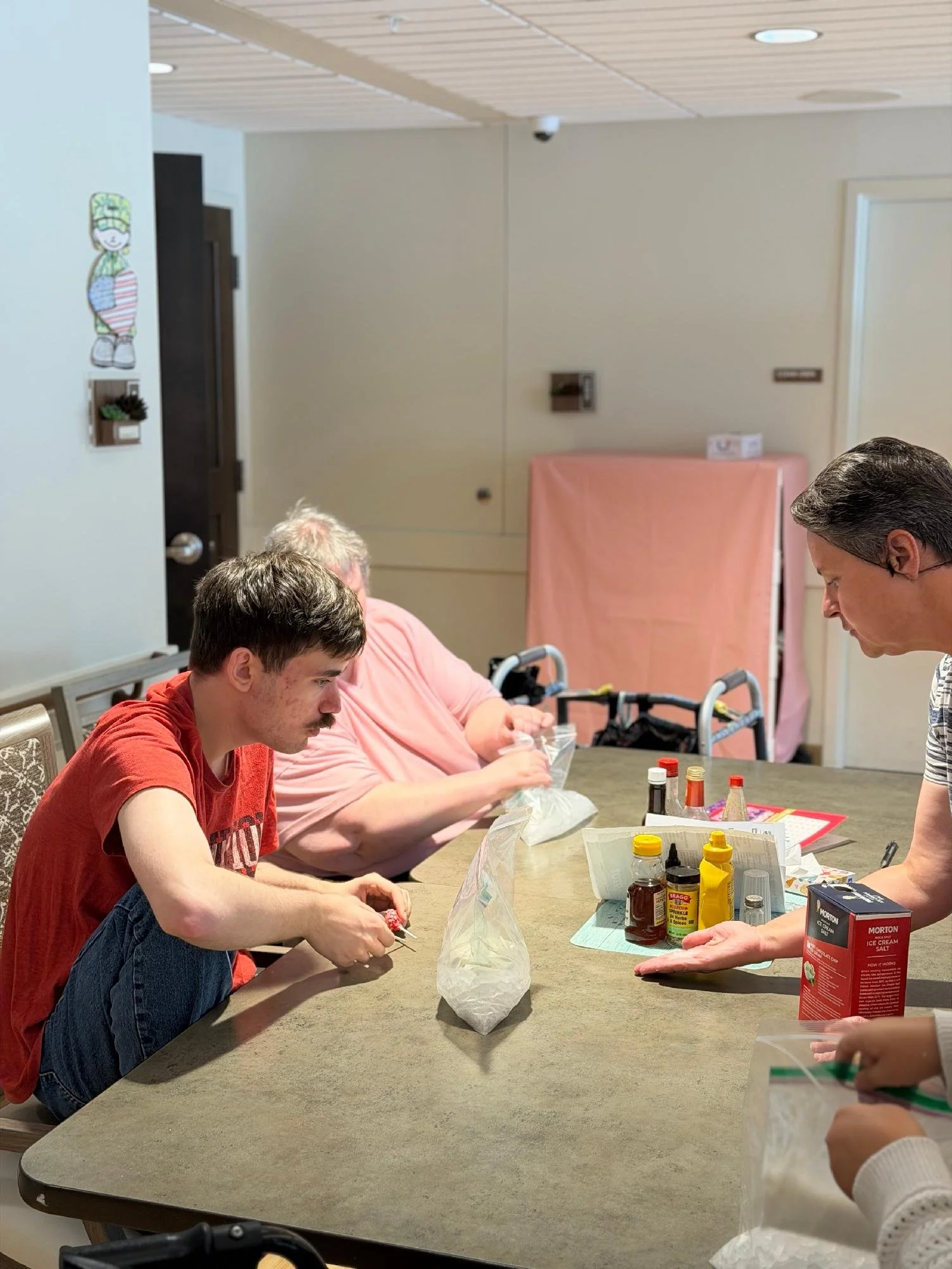 People sitting at a table working on a hands-on activity involving food items and condiments. There are various bottles and packages on the table, and the setting appears to be a community room.
