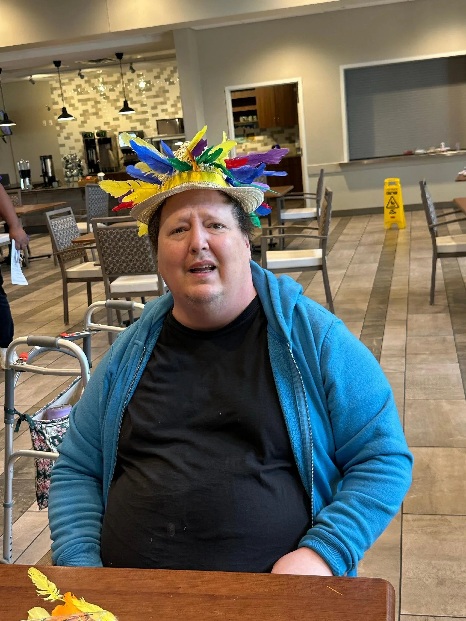 A man wearing a colorful feathered hat sitting in a restaurant or cafe with a cloudy glass of water, in a casual setting with tables and chairs.