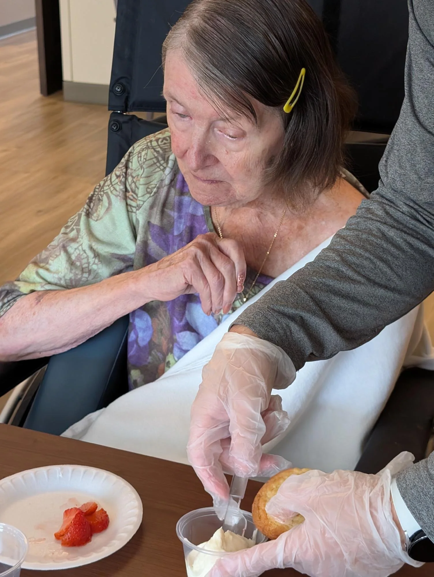 An elderly woman with gray hair held back with a yellow hairpin receiving care, possibly receiving food or medication, in a healthcare setting.