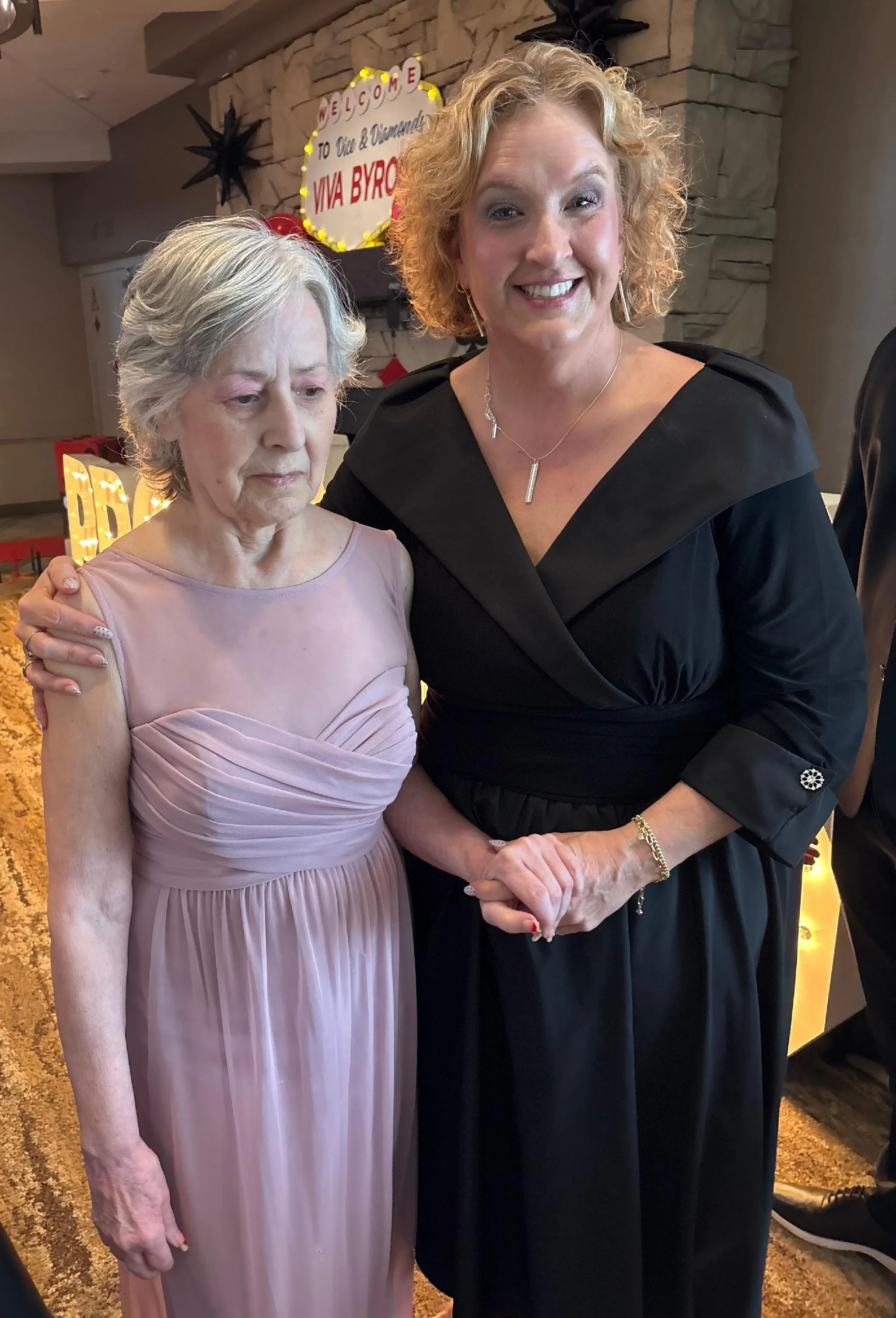 Two women standing together at an indoor event. One woman is elderly with gray hair wearing a pink dress, and the other woman has curly blonde hair wearing a black dress. Behind them is a sign saying 'Welcome to Ali & Cindy's Viva Byrd' with decorati