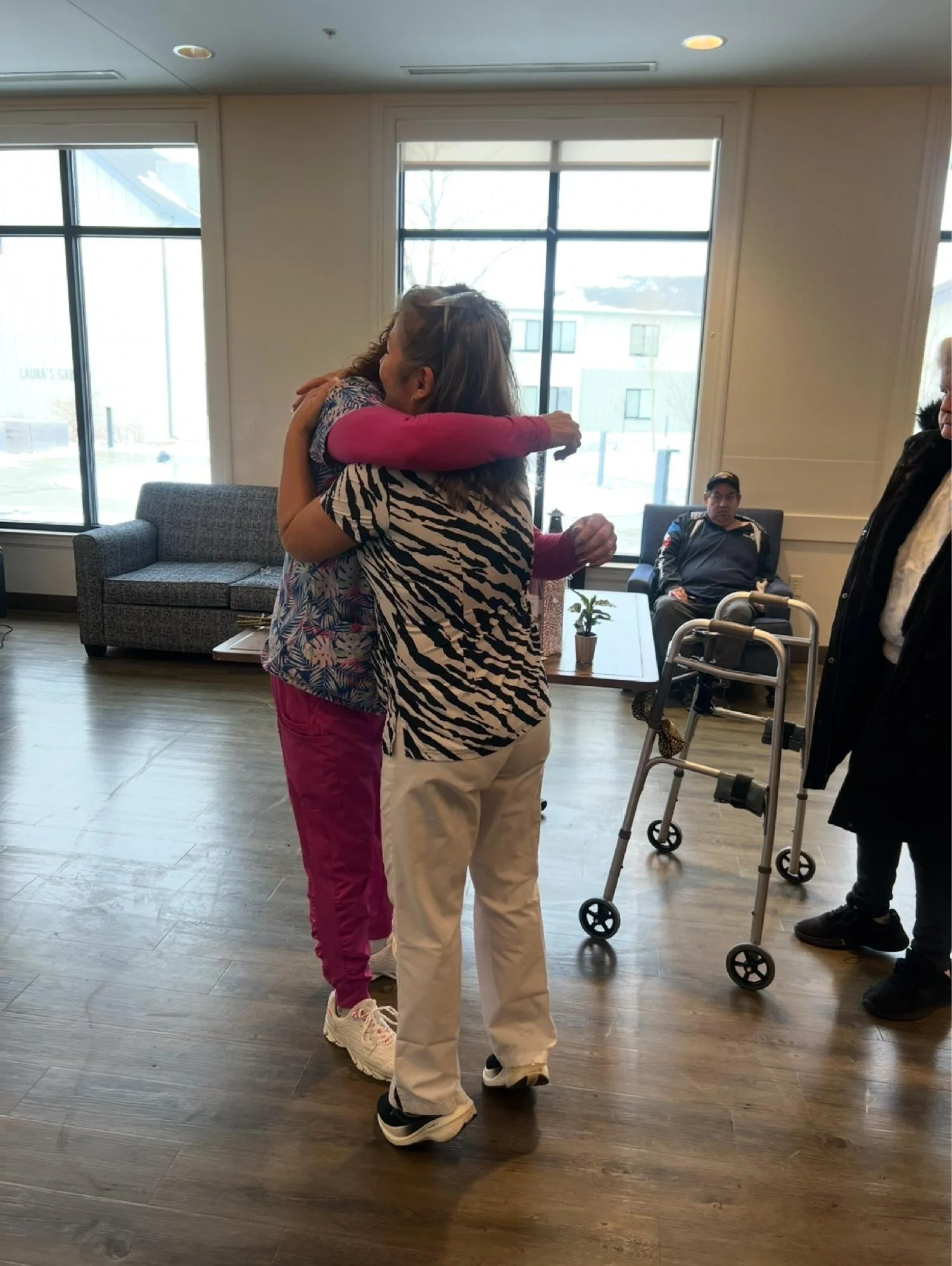 Two women hugging, one in a zebra-patterned top and the other in pink pants, stay in a room with large windows. In the background, a seated man and a woman with a walker are visible.