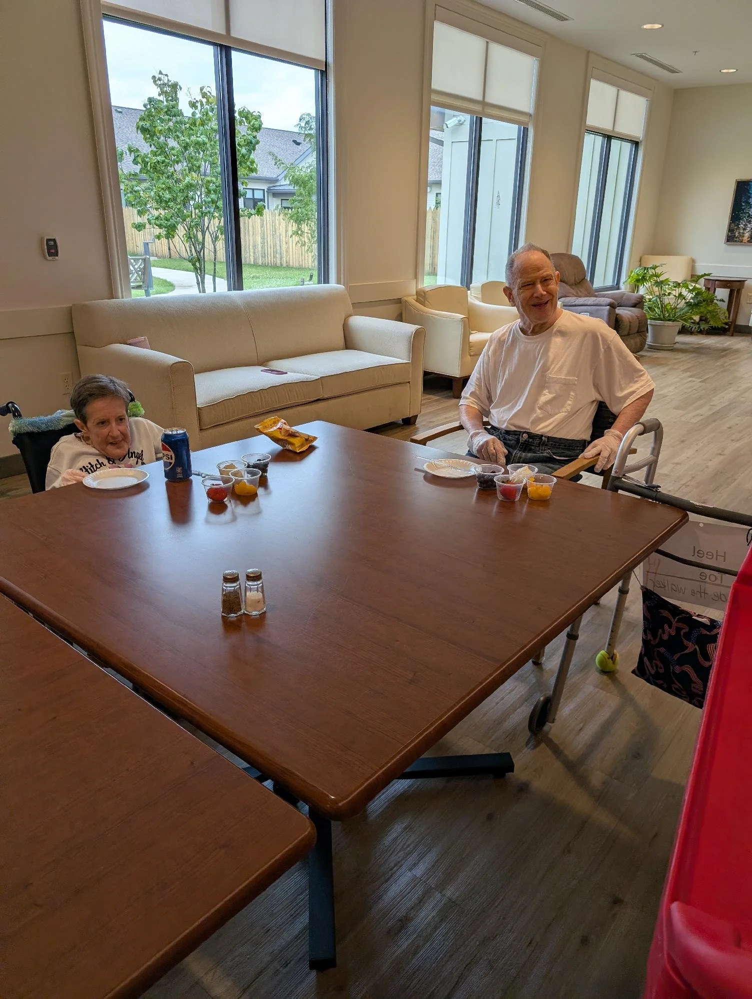 Indoor room with large windows, beige couches, potted plants, a wooden table with snacks and drinks, and an elderly man in a white t-shirt sitting in a wheelchair.