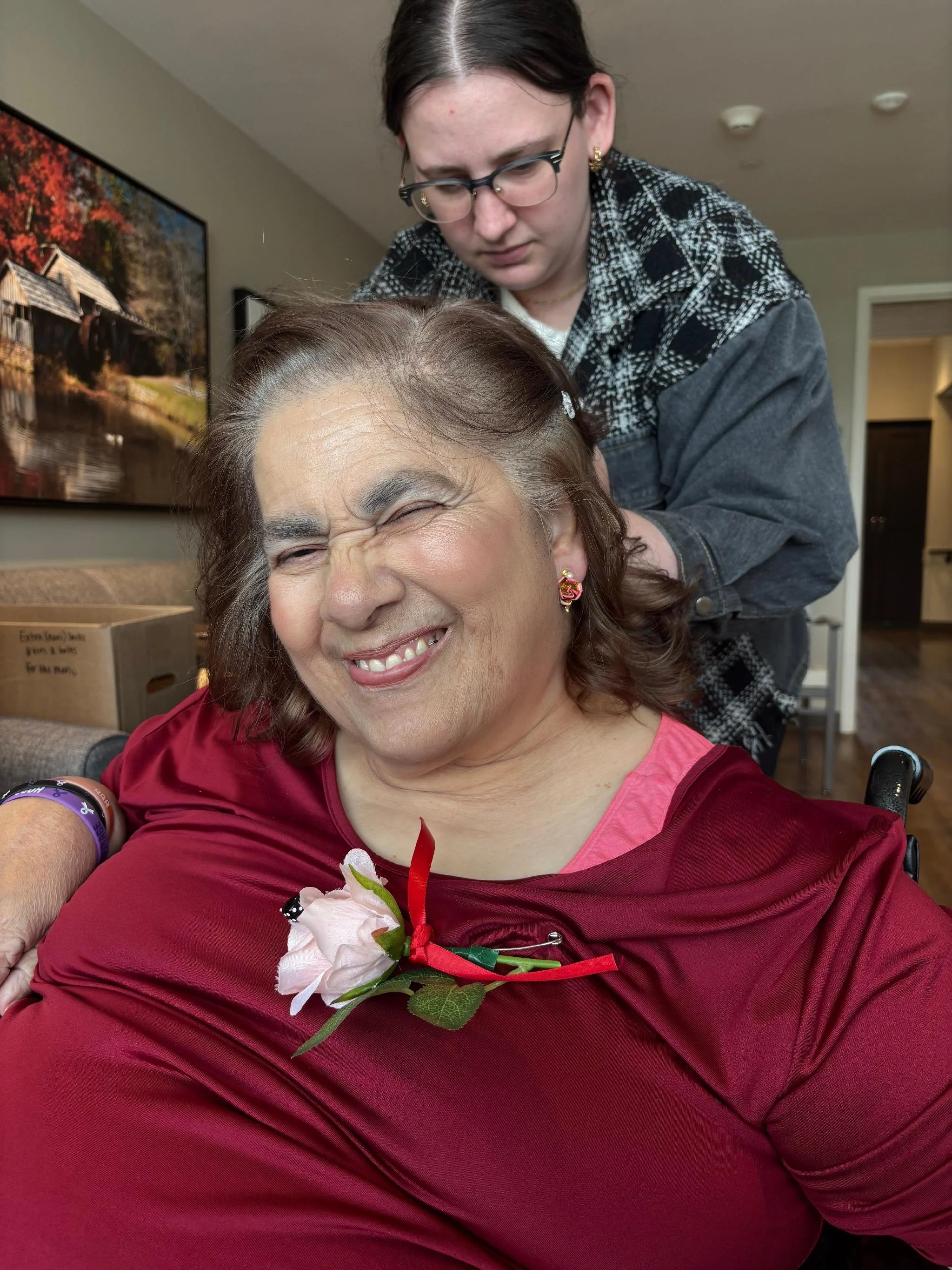 A woman with gray hair and earrings smiling with her eyes closed, wearing a pink shirt and a corsage, and a young woman with glasses and dark hair standing behind her, leaning over her in a room with beige walls, a painting of a house by a river, and