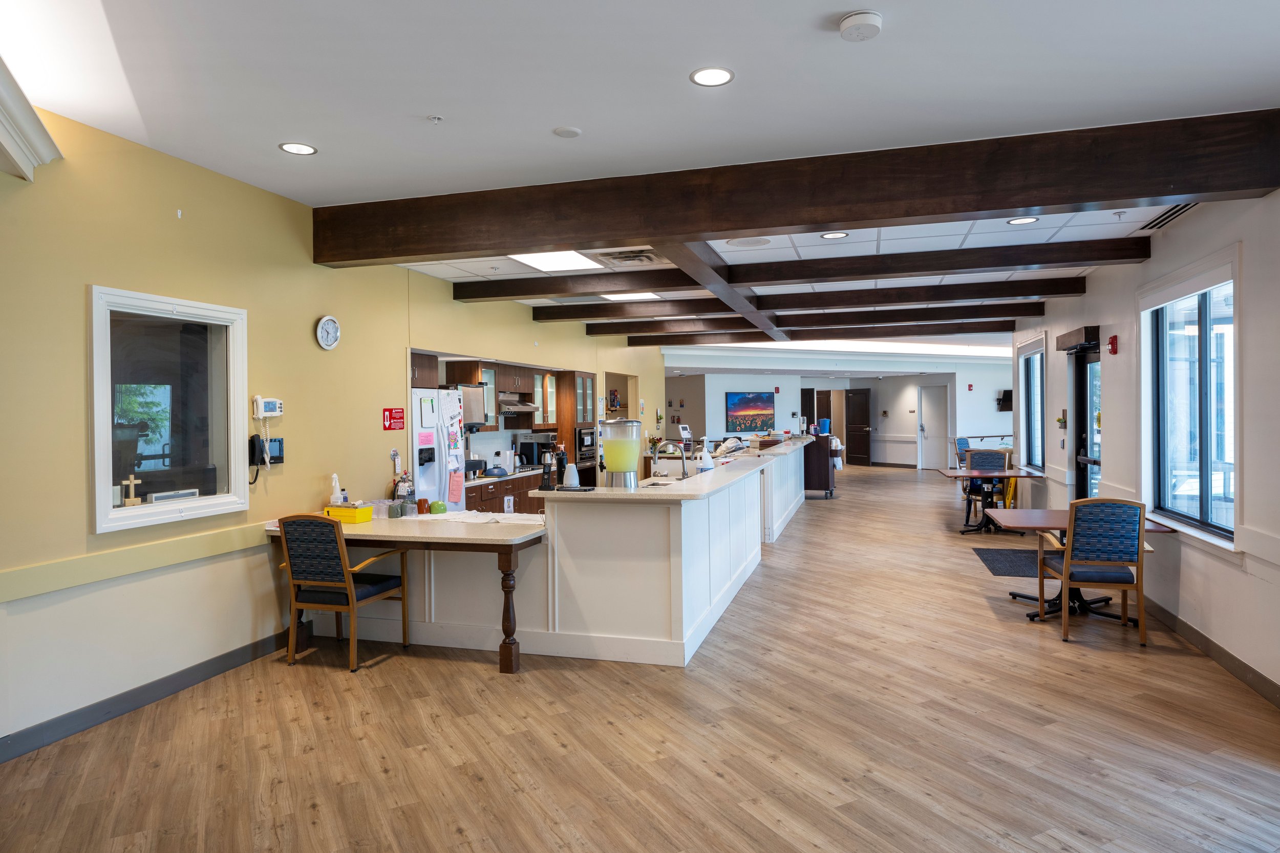 A spacious, well-lit common area with wooden flooring, a white counter, wooden beams on the ceiling, large windows, chairs, and a kitchen area with appliances.