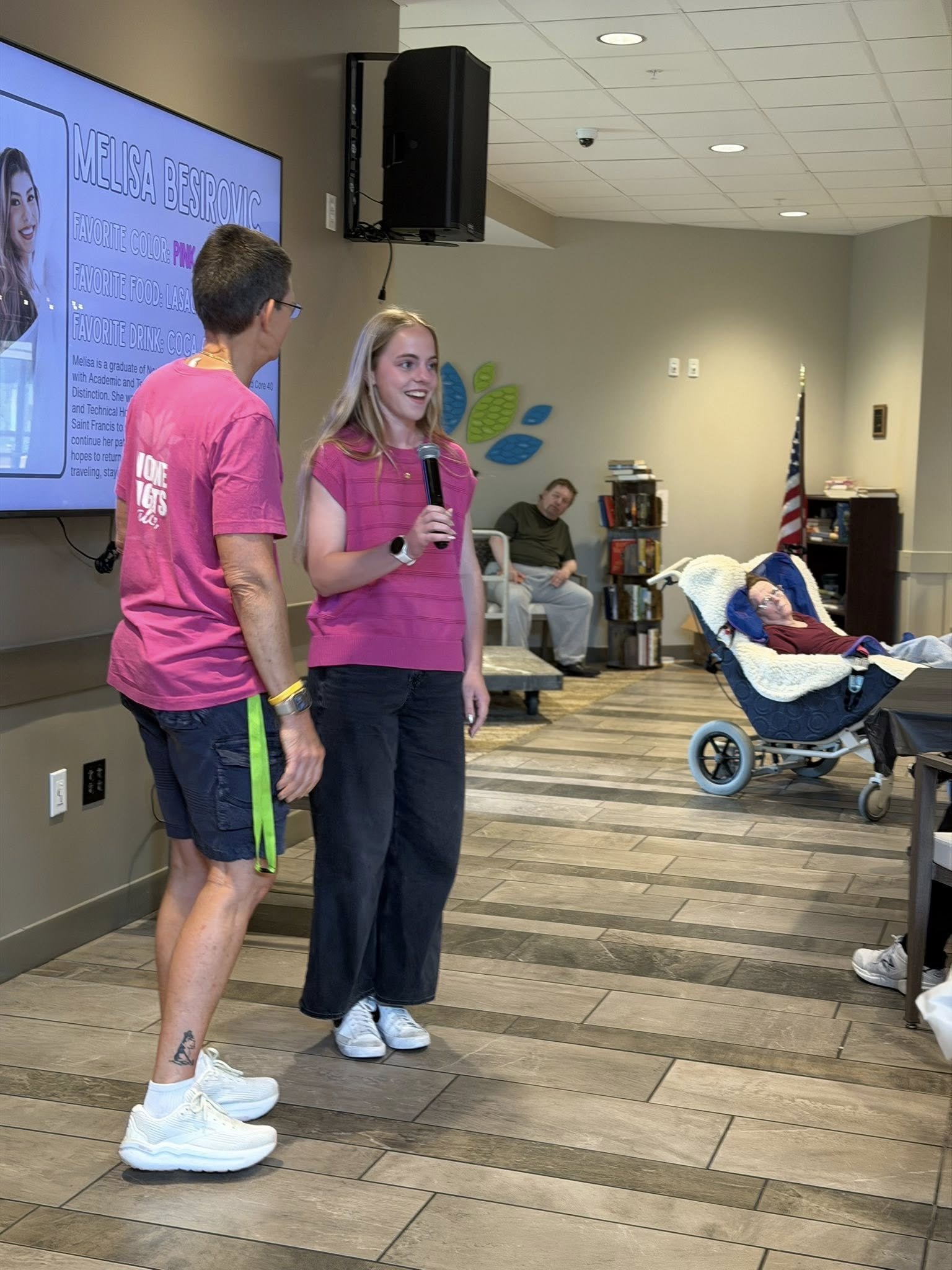 Two women standing in a room, one holding a microphone and both engaging in conversation. A person is lying in a stroller nearby. In the background, there is a large screen displaying information about Melissa Bejovic, including her picture, favorite