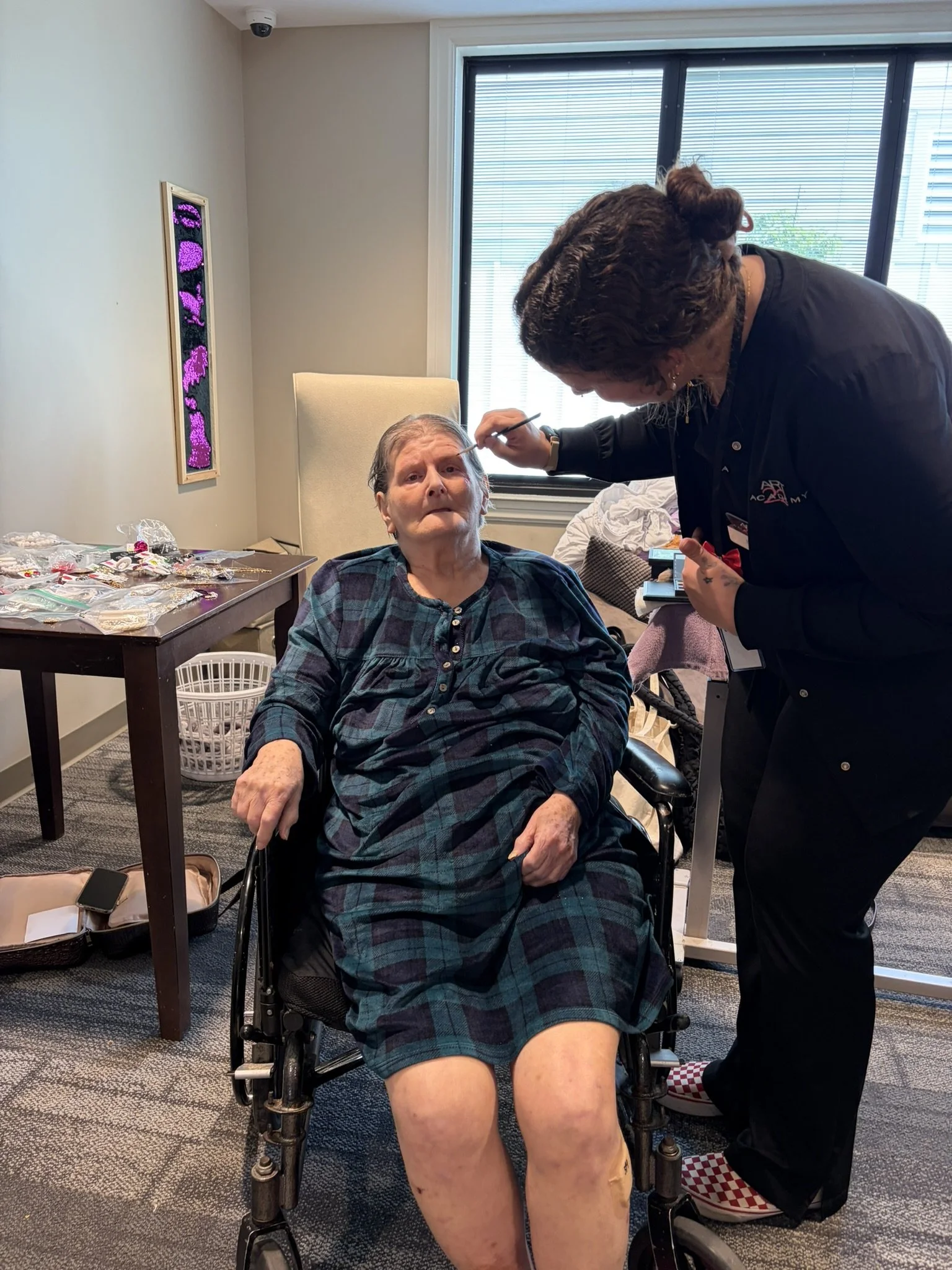 A woman in a plaid dress sitting in a mobility wheelchair getting her eyebrows done by a technician in a black shirt. The room has a window with blinds, a digital art piece on the wall, and a table with jewelry and other small objects.