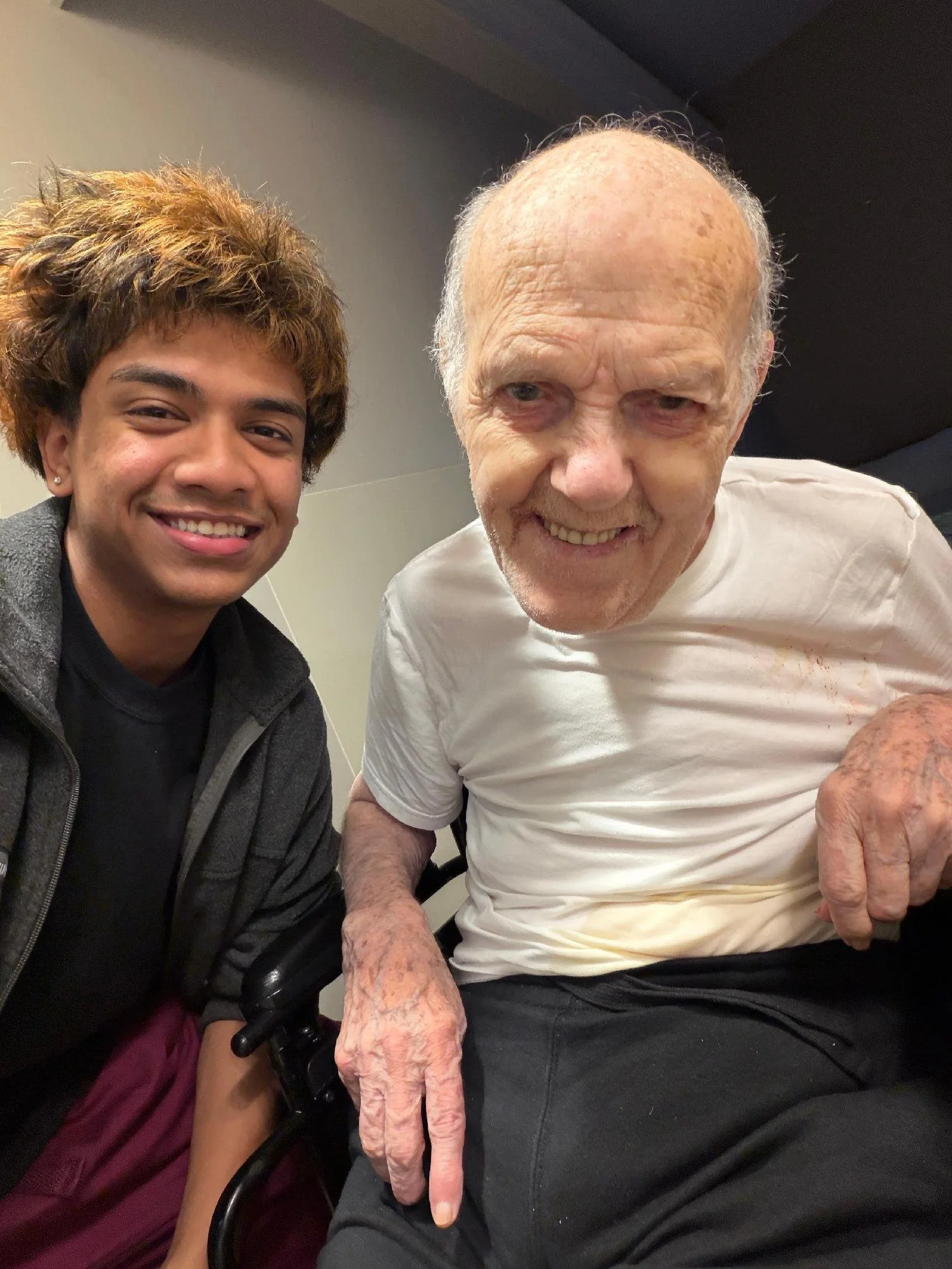 A young man with brown and blonde curly hair smiling for a selfie with an elderly man with a bald head, shiny skin, and a white shirt. The elderly man is showing his long middle finger.
