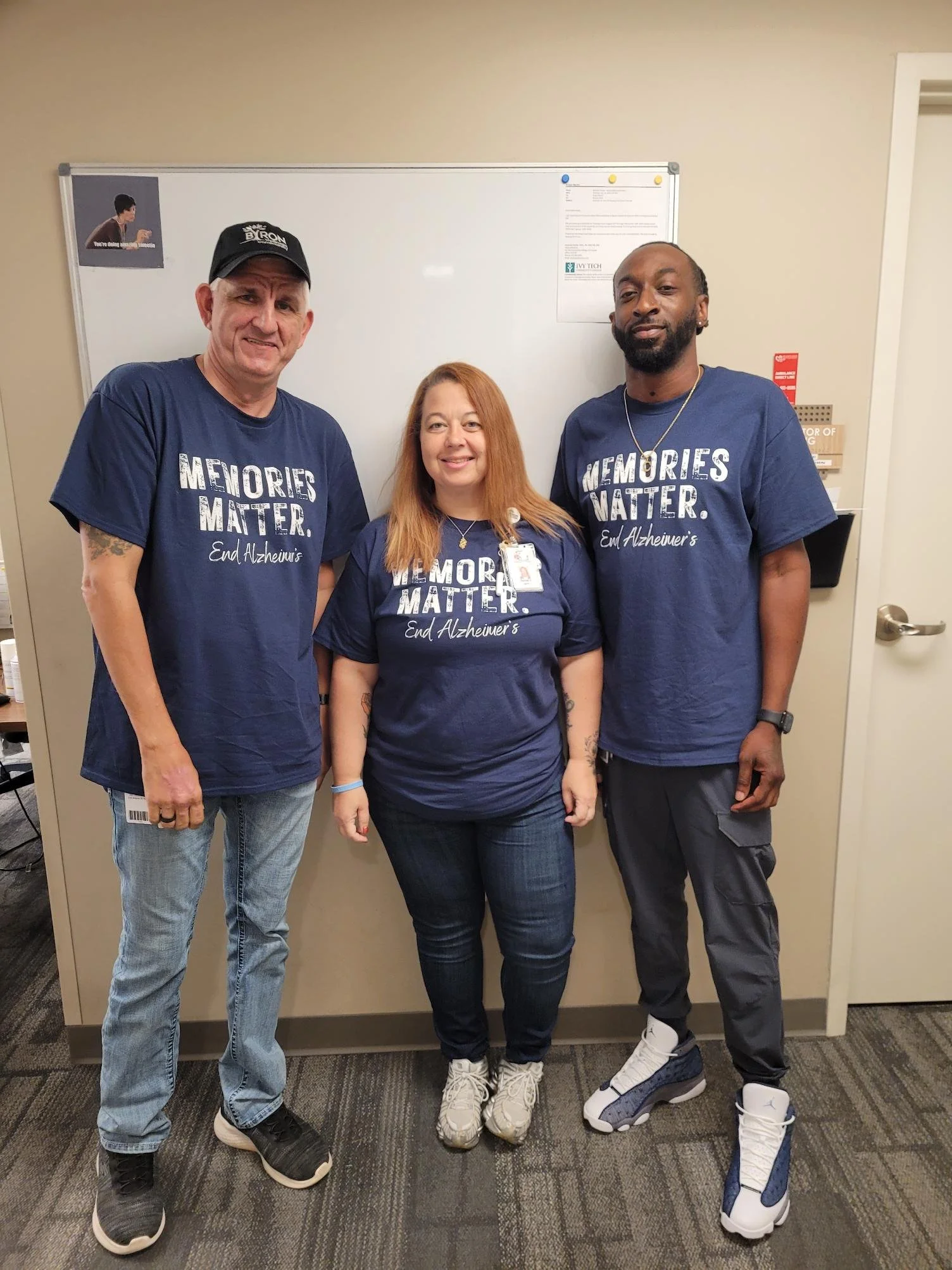 Three people standing in front of a whiteboard, wearing matching blue shirts with the text "memories matter, end Alzheimer's." The man on the left has a black cap, the woman in the middle has long red hair, and the man on the right has a beard and ea