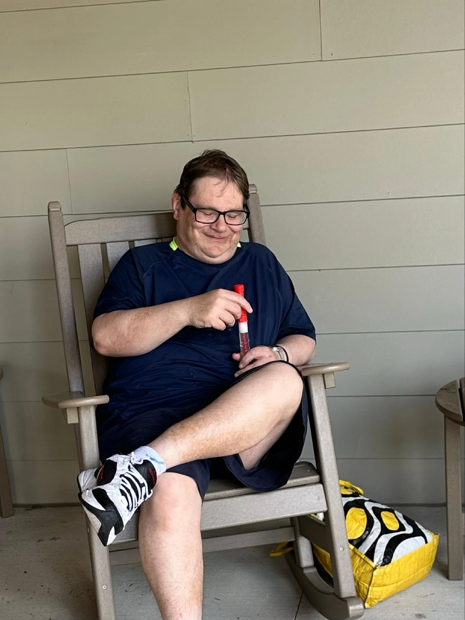 A man wearing glasses, a navy blue shirt, and black shorts sits in a beige wooden rocking chair. He is holding and looking at a red and white object, smiling. On the ground beside the chair, there is a yellow and black bag. The background has beige h