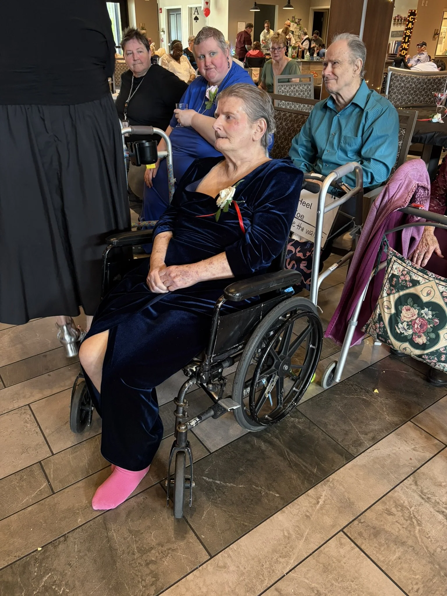 A group of elderly people, some in wheelchairs, gathering indoors at a social event. The focus is on a woman in a black dress with a white flower and a limp hand, seated with a pink sock on her foot, and a man in a teal shirt next to her. Other indiv