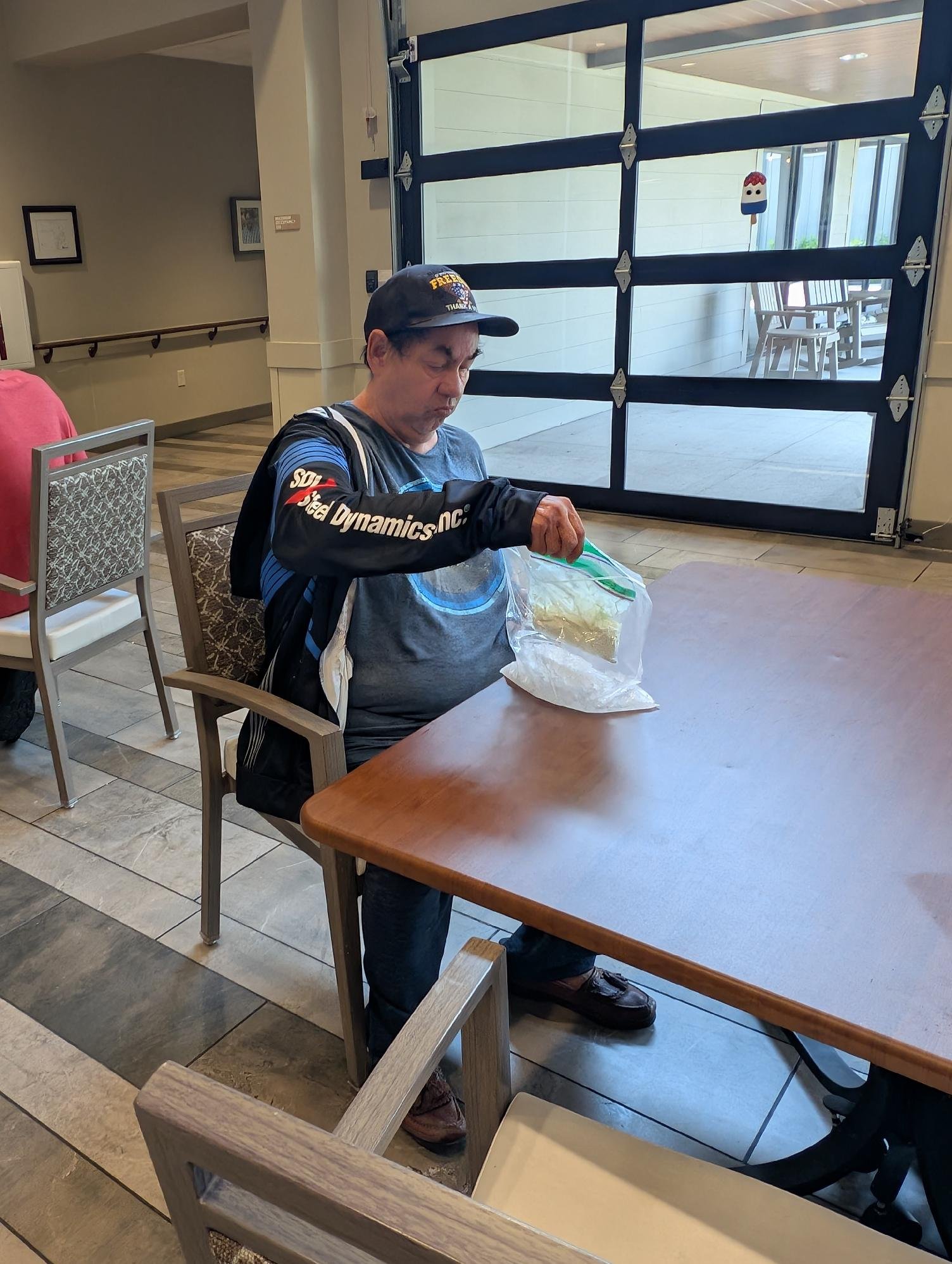 A man seated at a wooden table in a restaurant or cafe, looking into a plastic bag, wearing a dark jacket with text on the sleeve and a baseball cap, near a large garage-style window with outdoor patio furniture visible outside.
