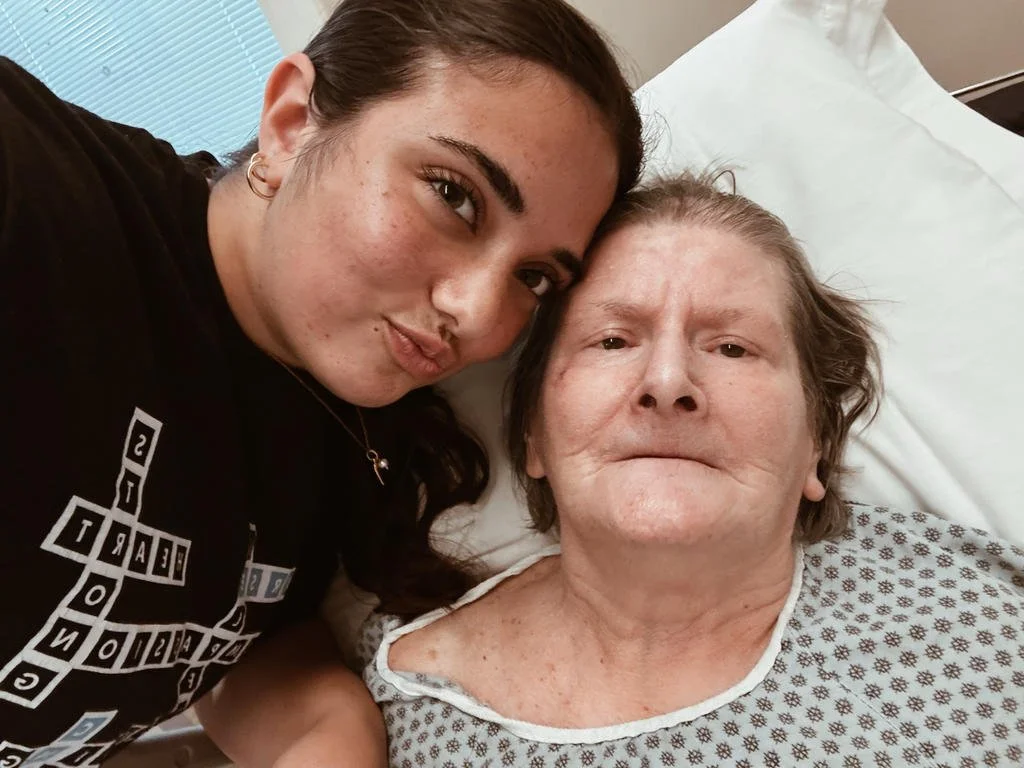 Two women taking a selfie in a hospital room, one young and one older, lying in bed, both looking at the camera.