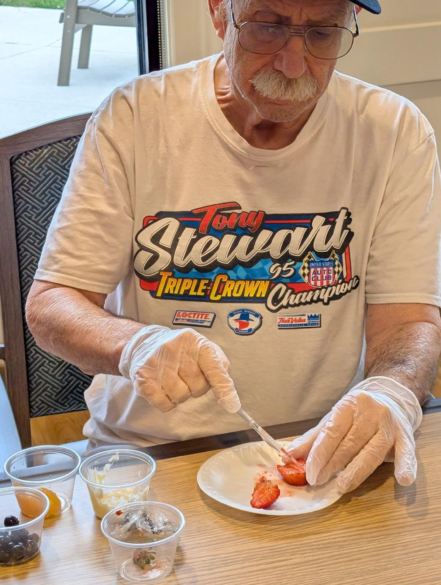 An elderly man with a mustache, wearing glasses and a baseball cap, is preparing strawberries with a syringe. He is seated at a wooden table with small containers of various fruit sauces or toppings nearby. He is wearing a white T-shirt with racing-t