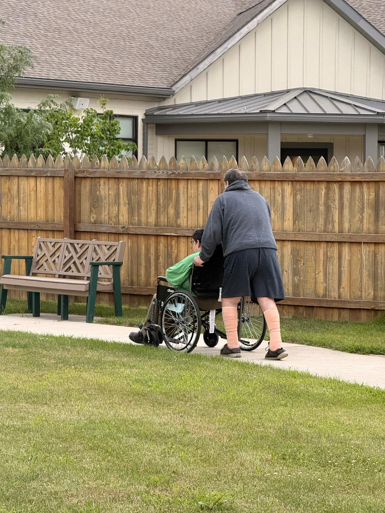 Person in gray sweater and woman in pink leg warmers pushing a person in a wheelchair along a sidewalk in a backyard with a wooden fence and house in the background.