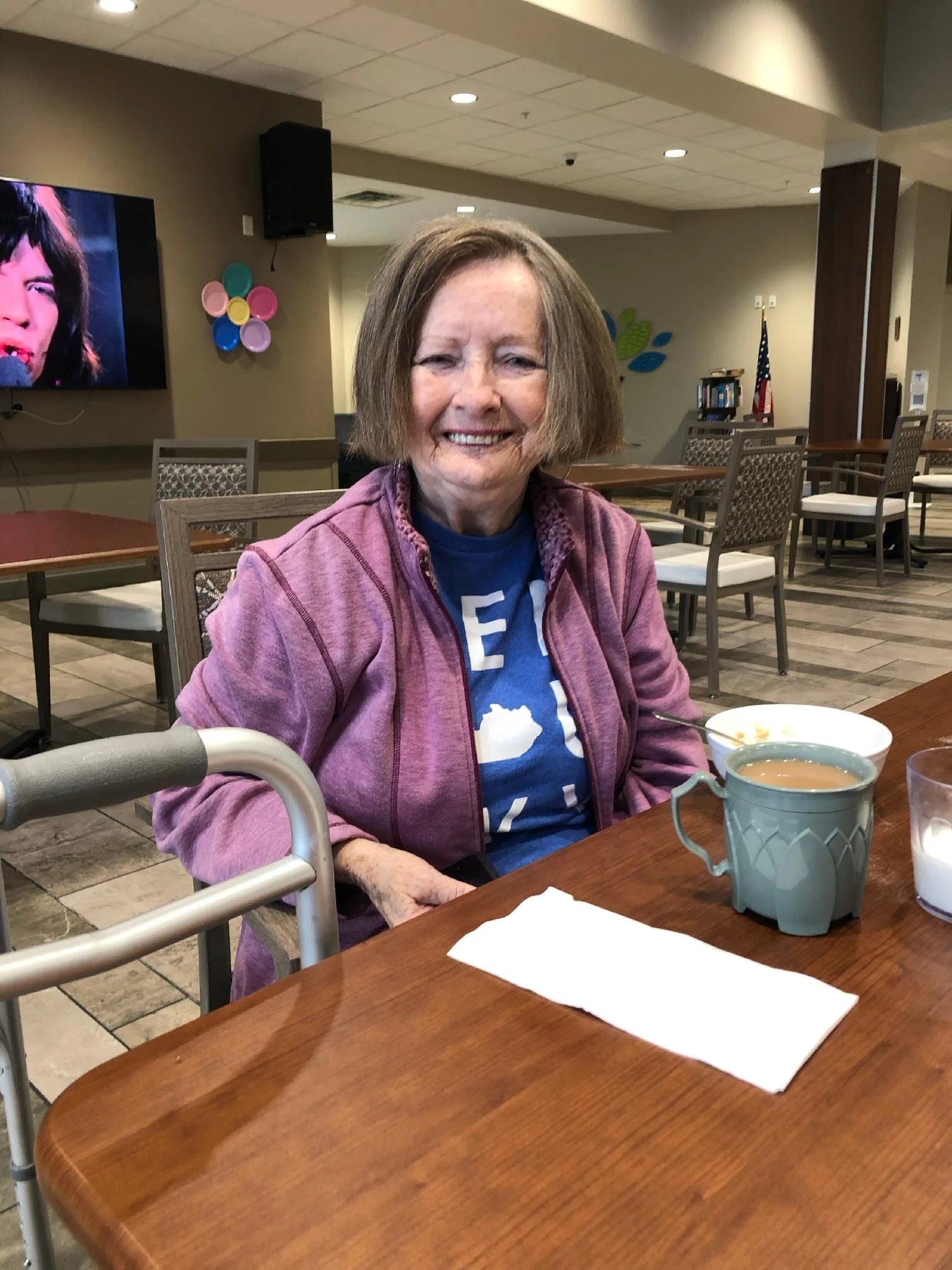 An elderly woman with shoulder-length brown hair, wearing a purple jacket and a blue shirt, sitting at a wooden table in a cafe or restaurant. She is smiling and has a cup of coffee in front of her. The background includes chairs, a television screen