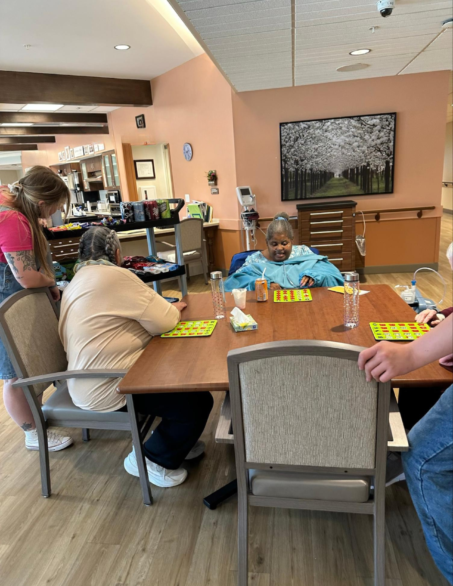 A nurse or caregiver sitting at a table with three elderly women, two of whom are playing bingo and the third preparing to play. The room has warm pink walls, wooden flooring, and a large flat-screen TV showing a scenic image of cherry blossoms.