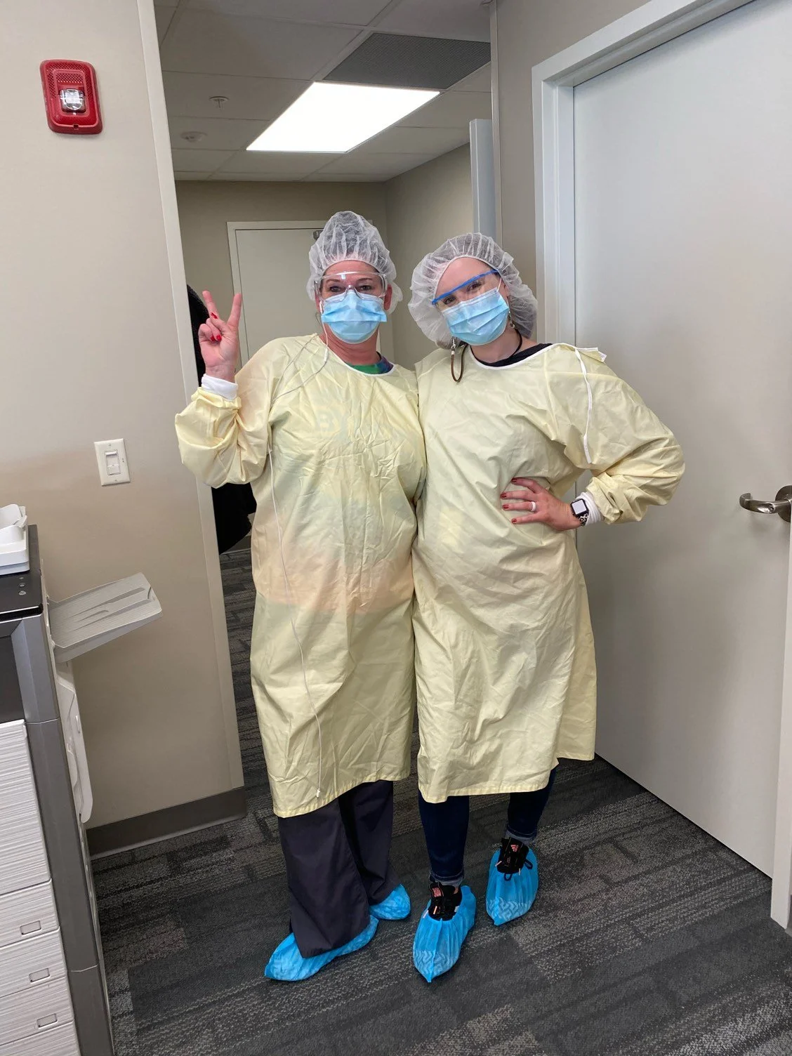 Two healthcare workers in yellow protective gowns, face masks, hair covers, and shoe covers, standing together in a hospital hallway, posing for a photo with one making a peace sign.