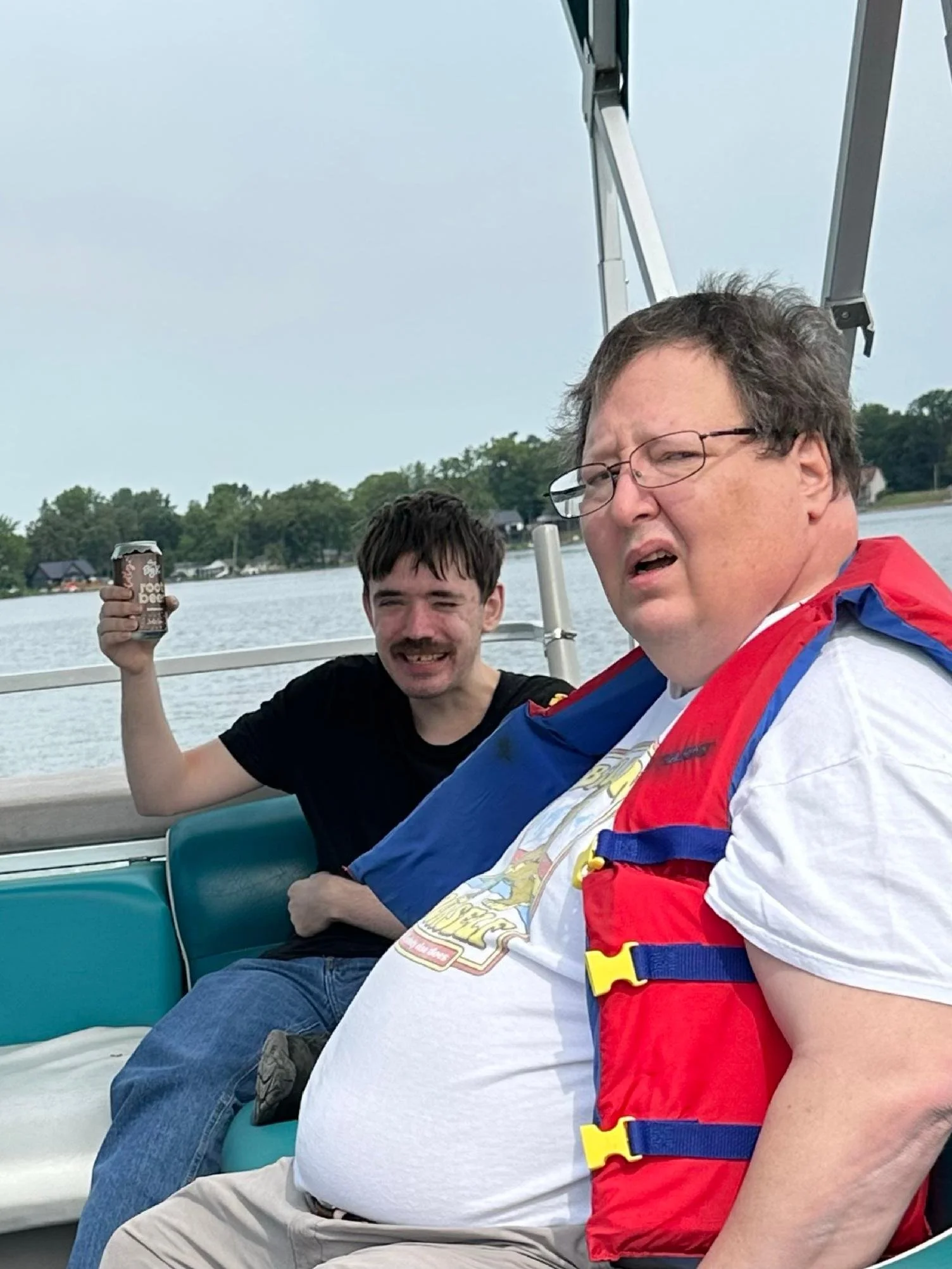 Two men sitting on a boat, one holding a can of soda, with water and trees in the background.