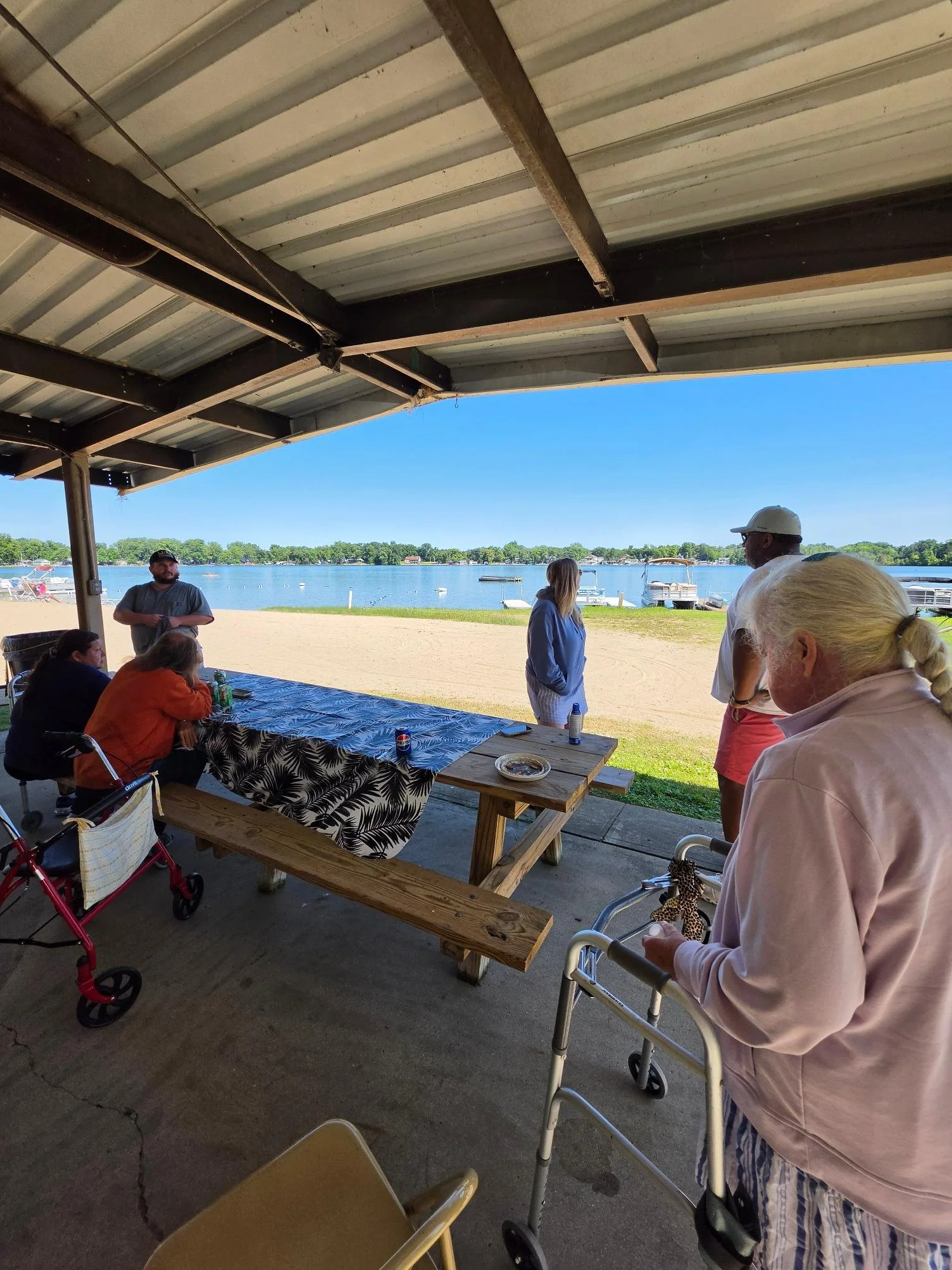 People gathered under a shelter by a lakeside, some sitting at a table, others standing, with boats docked on the water and trees in the background.