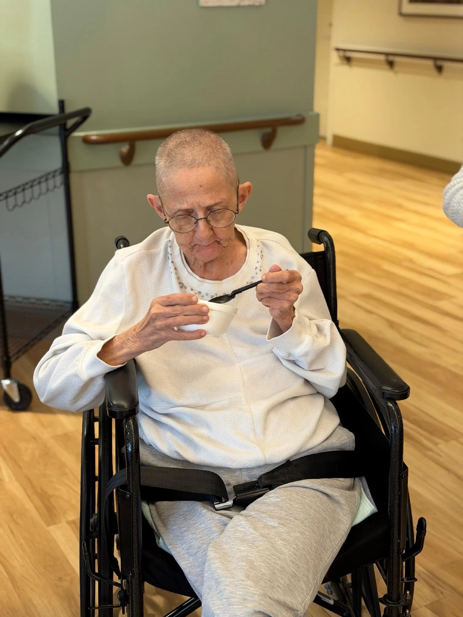 An elderly woman with short gray hair and glasses sitting in a wheelchair, eating from a white bowl with a black spoon in a care facility.