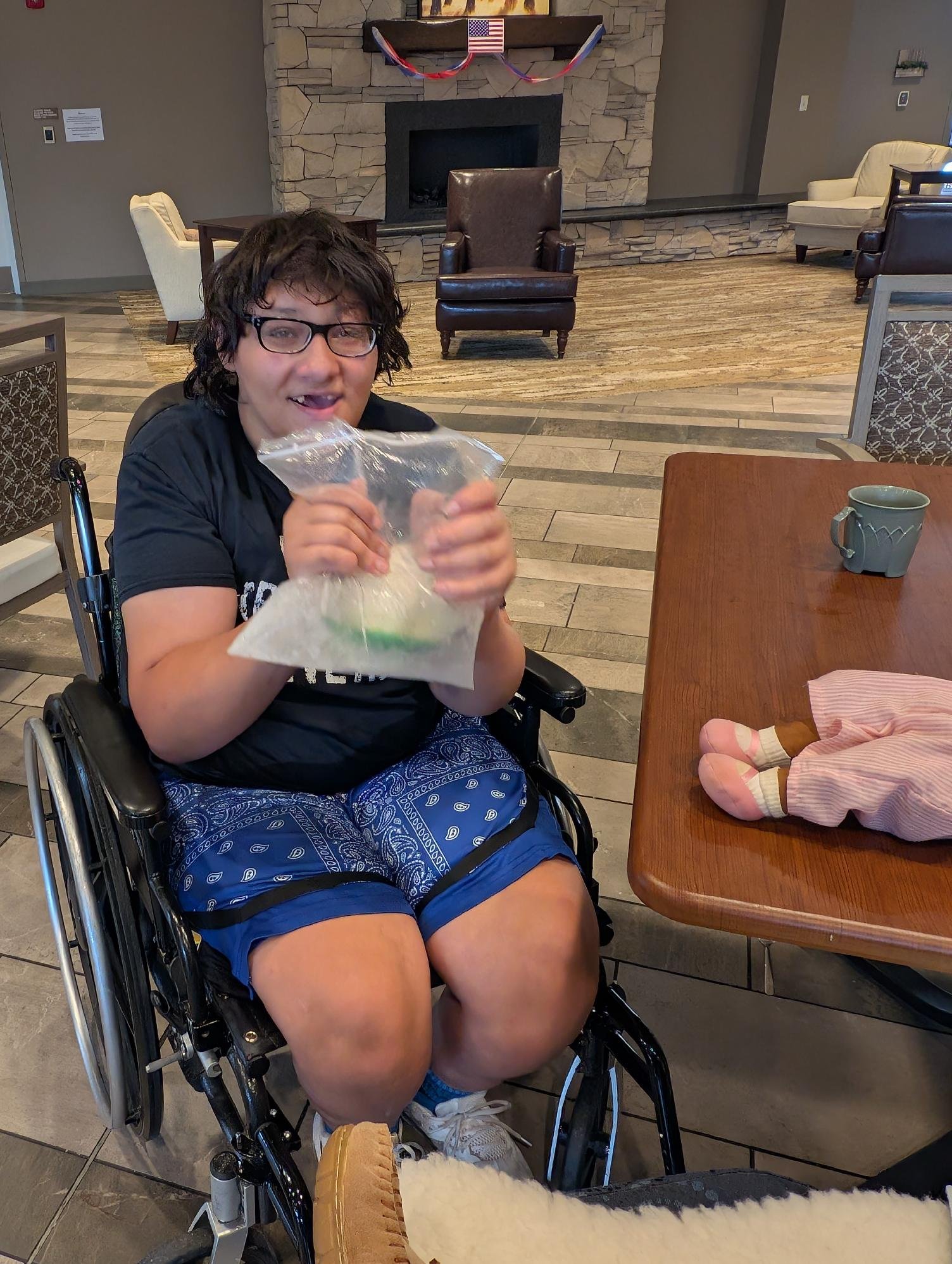 A young person with glasses, dark curly hair, and a black T-shirt sitting in a wheelchair. They are holding a plastic bag and smiling at the camera. In front of them on a wooden table, there is a pink and white striped plush doll wearing pink pajamas