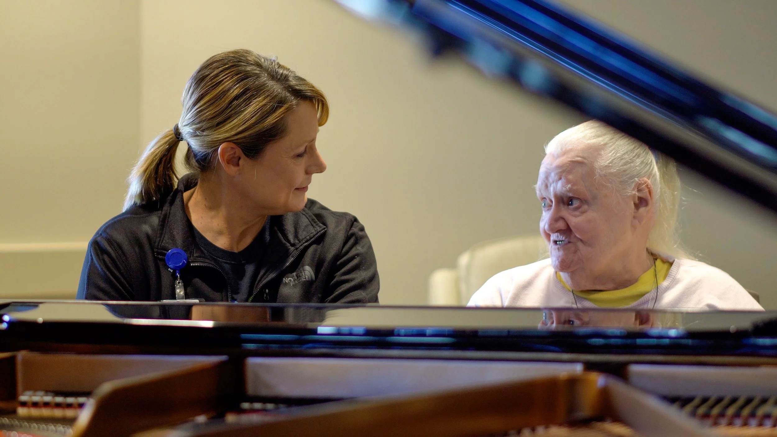 A caregiver talking to an elderly woman at a piano.