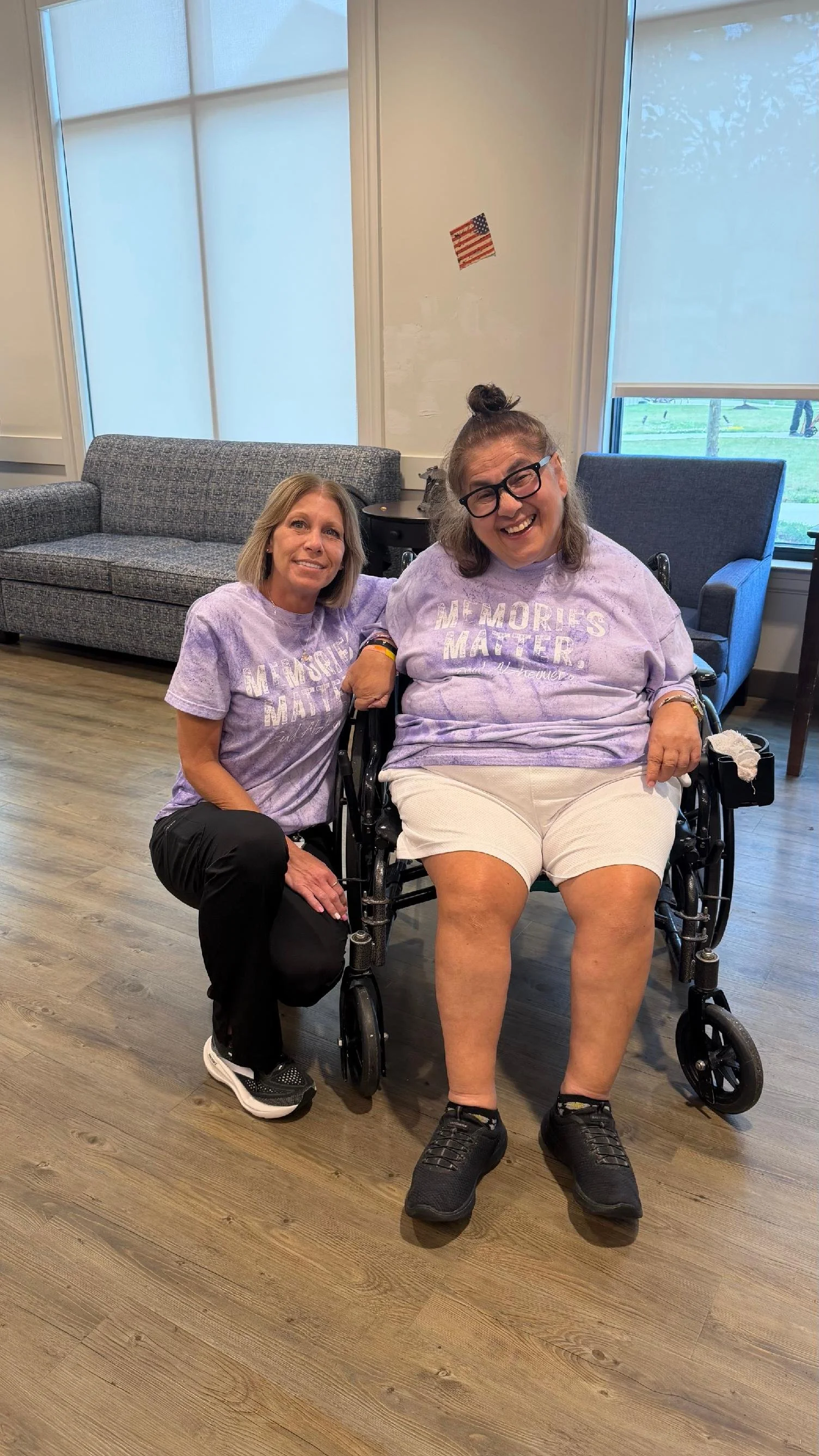 Two women, one sitting in a wheelchair and another kneeling beside her, both smiling. They are wearing matching purple shirts with the phrase 'Memories Matter' on them. The room has large windows, a gray couch, and an American flag sticker on the wal