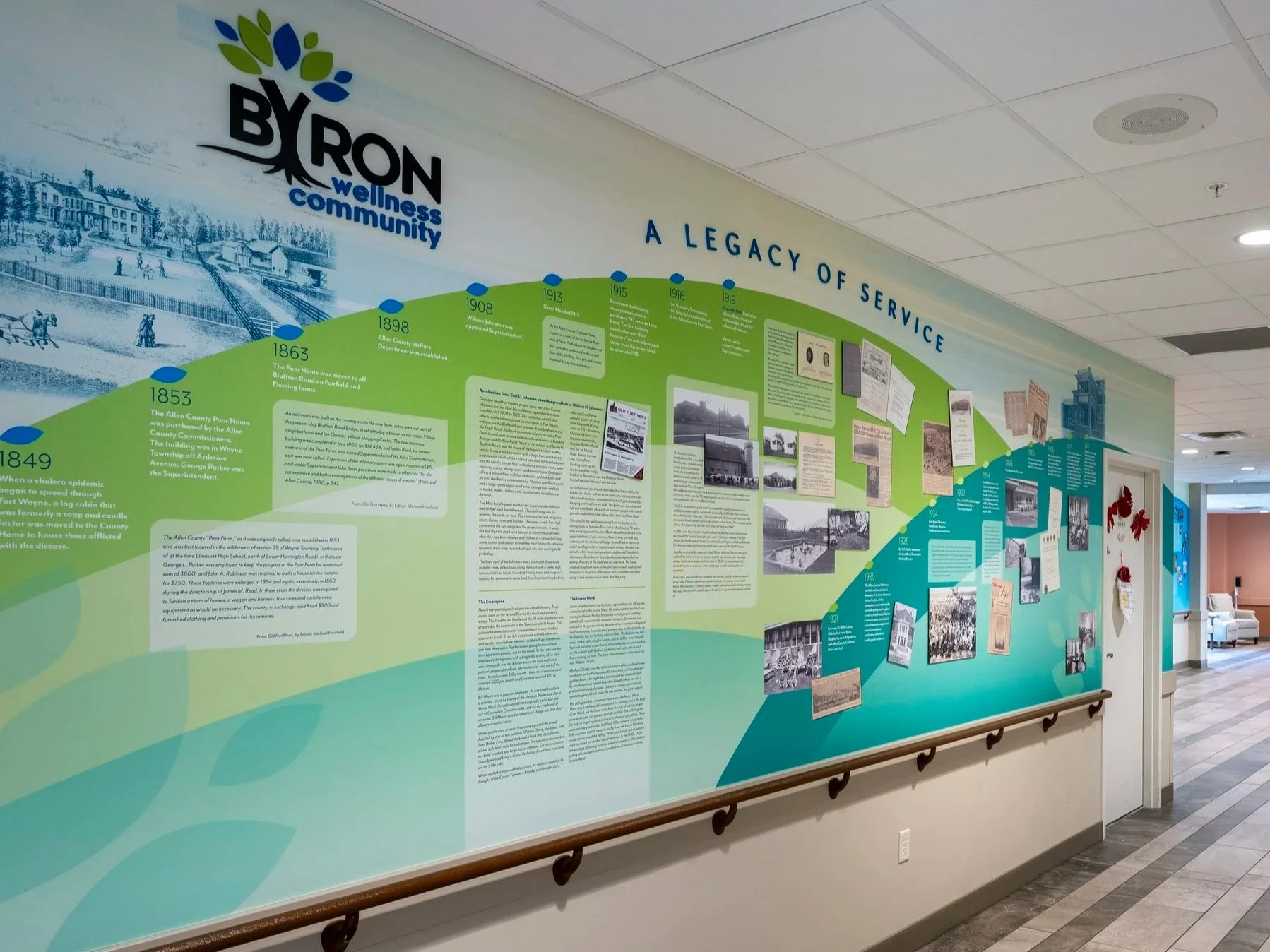 A large indoor display wall titled "A Legacy of Service" with historical information, photographs, and documents related to the history of Byron, a wellness community. The wall features a timeline starting from 1849 up to the 1900s, highlighting key events in the community's history, and is decorated with a logo that includes stylized leaves and the words "BYRON wellness community." The hallway has ceiling tiles, a fire alarm, and a seating area nearby.