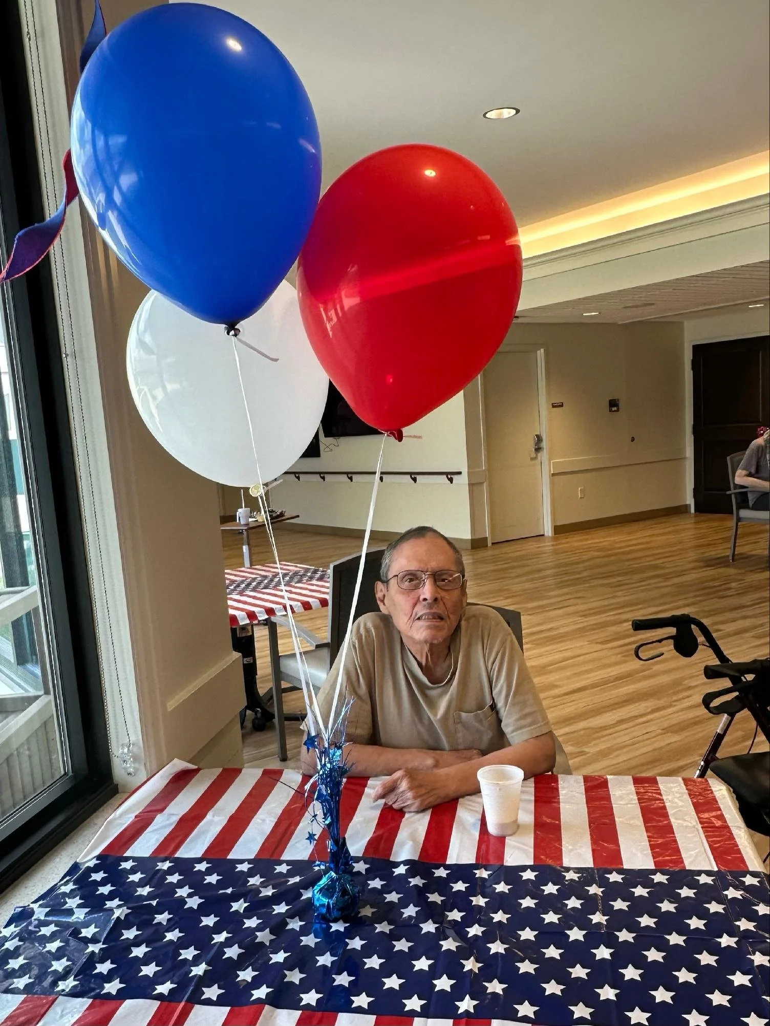 An elderly man sitting at a decorated table with patriotic red, white, and blue tablecloth, surrounded by balloons in red, blue, and white, celebrating a patriotic event in a bright room.