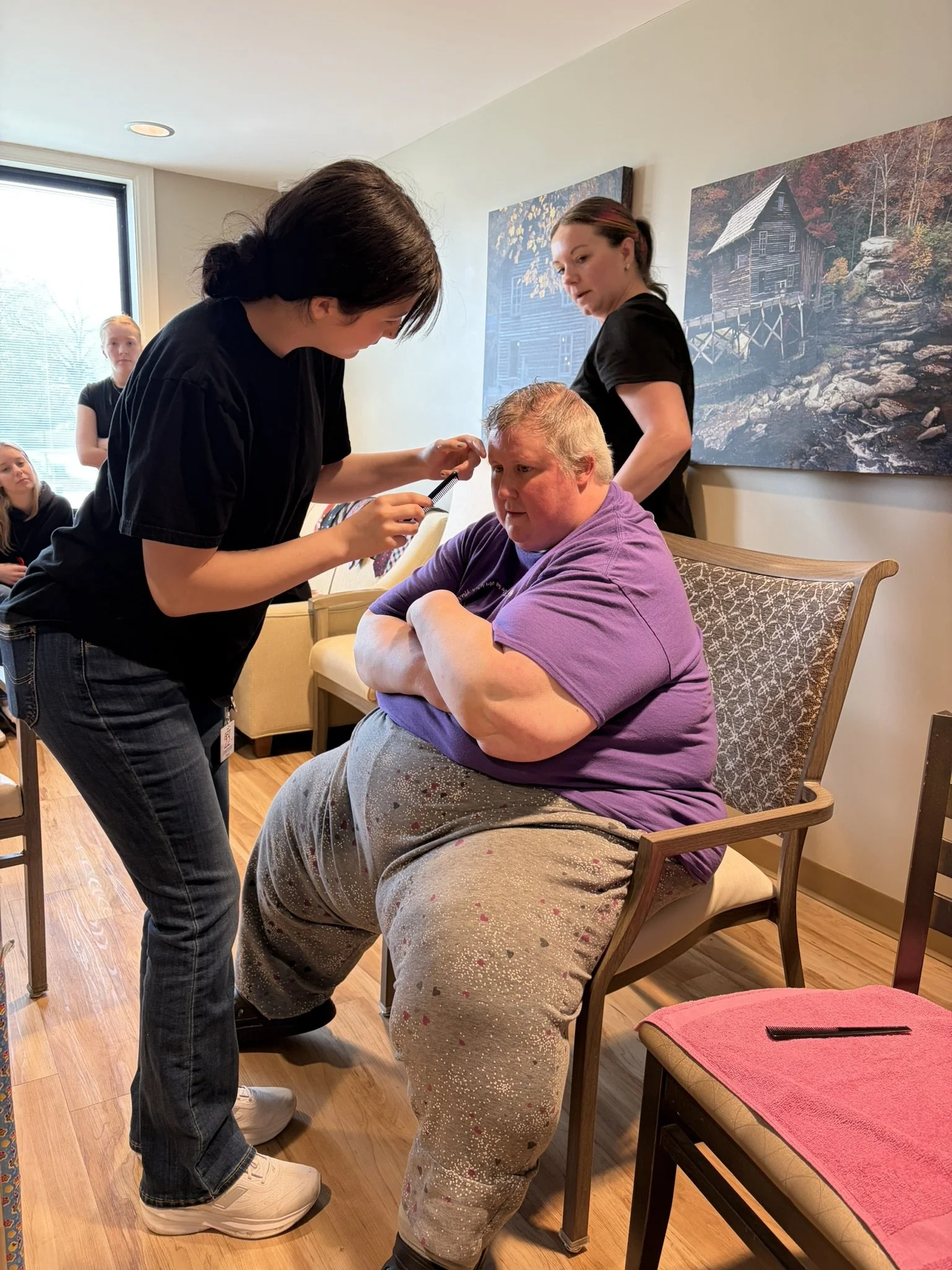 A person is getting a haircut from a barber in a cozy salon while three women sit nearby observing. The person getting the haircut is sitting in a chair with arms crossed, wearing a purple shirt and patterned pants. The barber is focused on cutting h