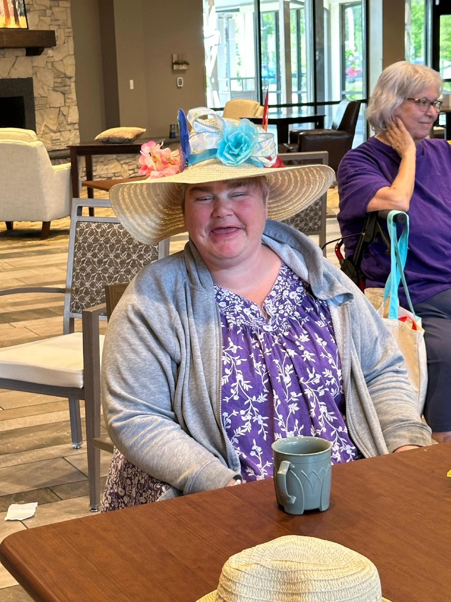 A woman sitting at a restaurant table wearing a large straw hat decorated with colorful artificial flowers and ribbons. She is smiling with her eyes closed, holding a mug, and dressed in a purple patterned dress with a gray hoodie. Another woman, dre
